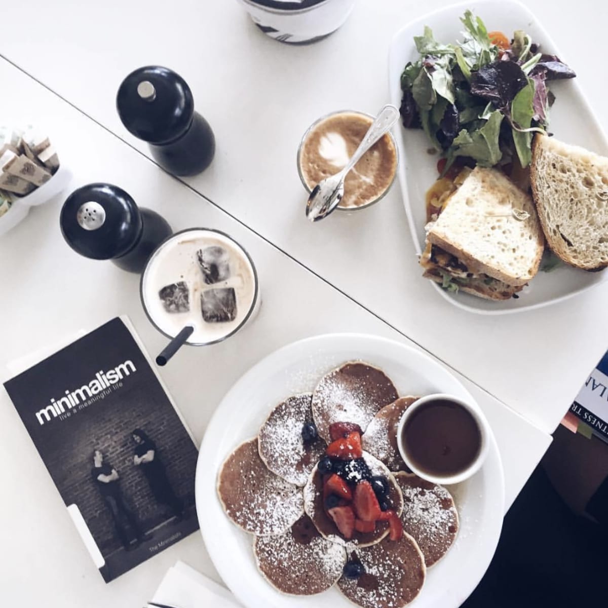A table with a plate of pancakes topped with strawberries and syrup, a sandwich with salad on the side, and a coffee drink, along with a minimalist magazine.