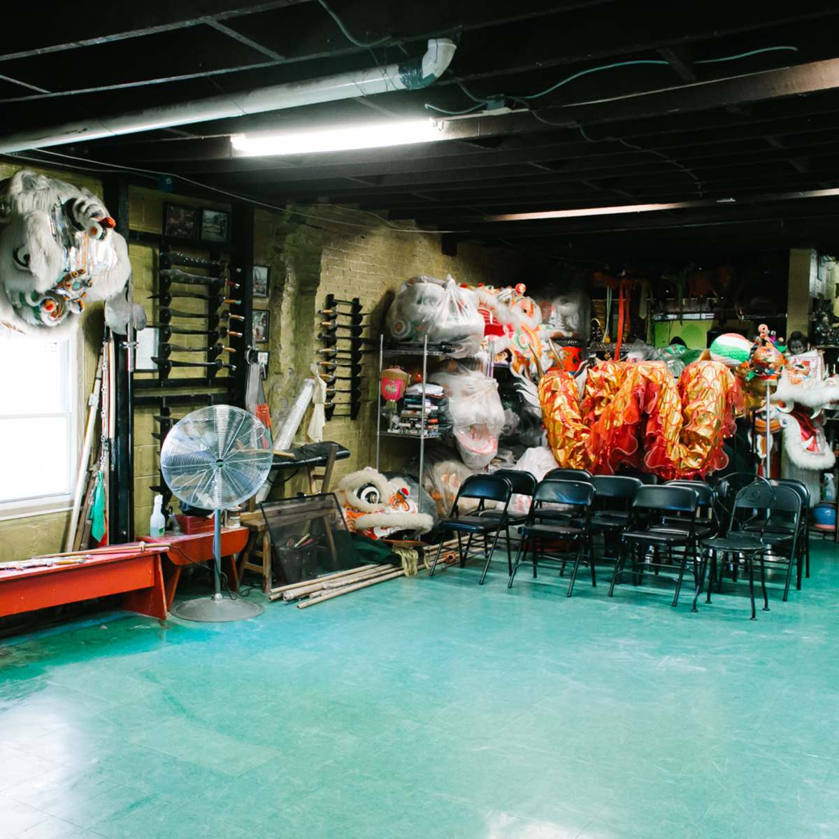 An indoor space with martial arts weapons on the walls, a fan in the corner, and colorful dragon and lion costumes hanging next to empty chairs.