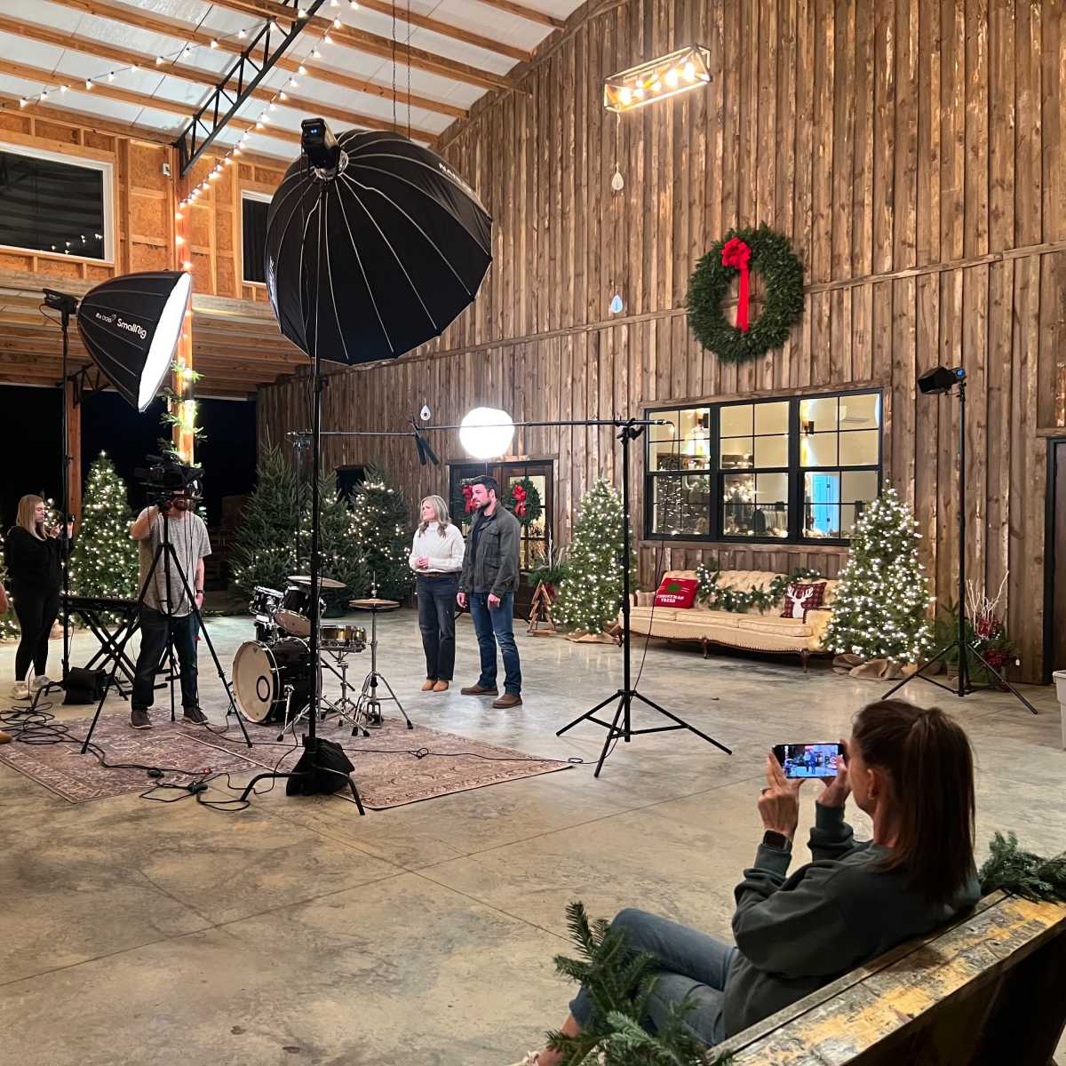 A group of people gathers around a photography setup inside a decorated barn with Christmas decorations and studio lighting equipment.