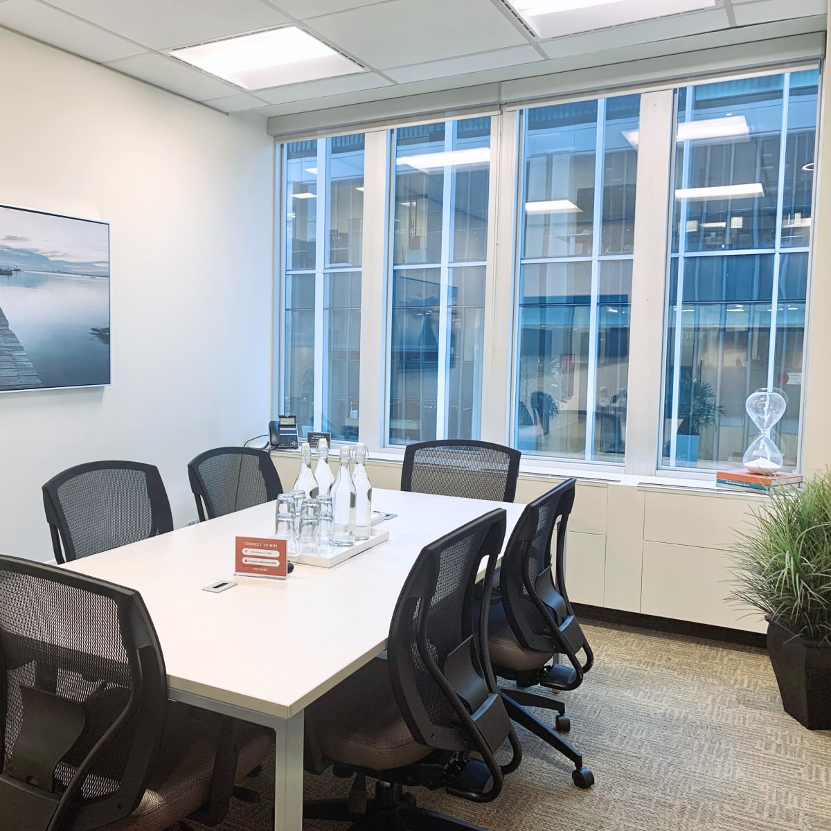 A conference room features a large white table surrounded by ergonomic chairs, with a scenic photograph on the wall and large windows allowing natural light.
