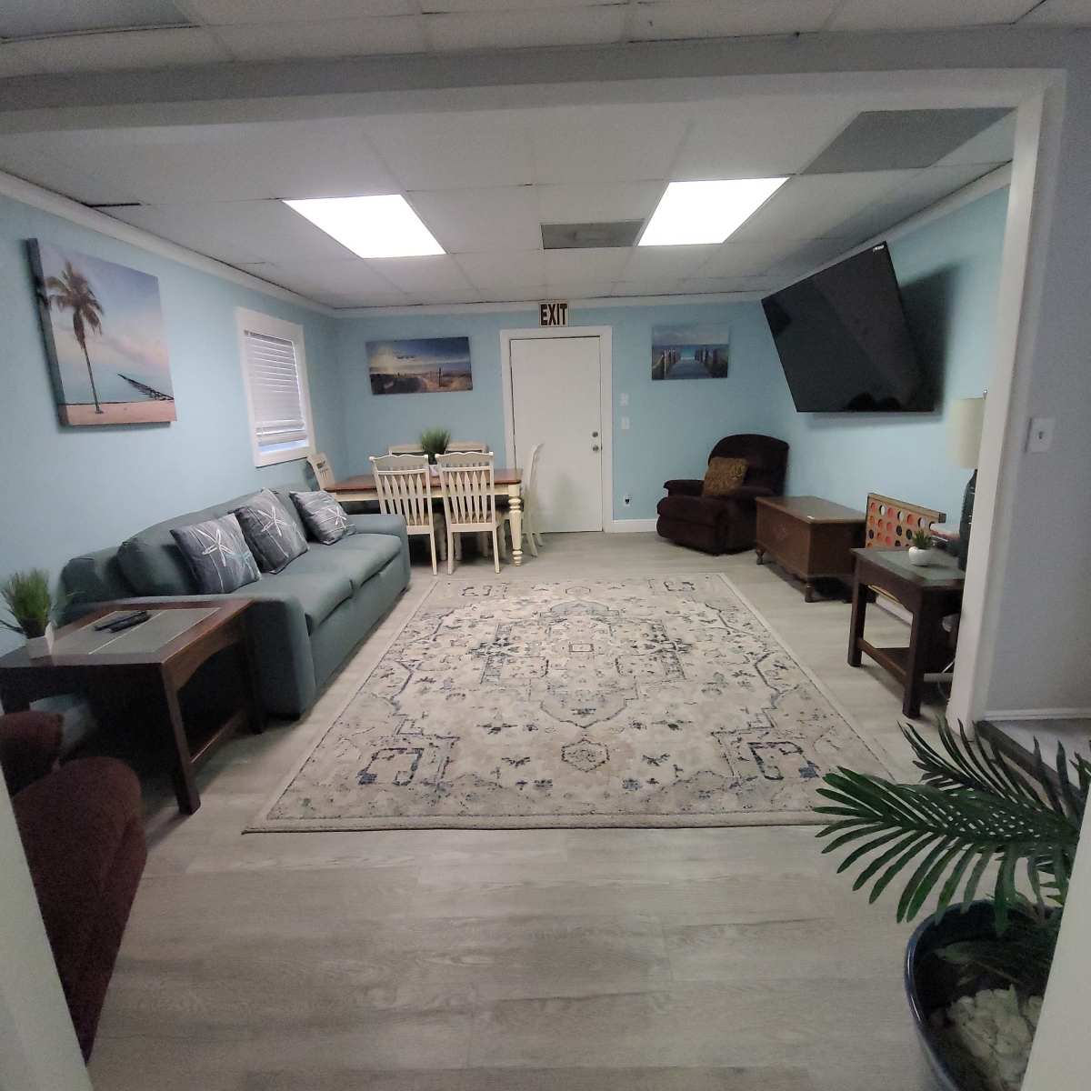 A light blue living room features a sofa, armchair, dining table, and a large rug, with a television mounted on the wall.