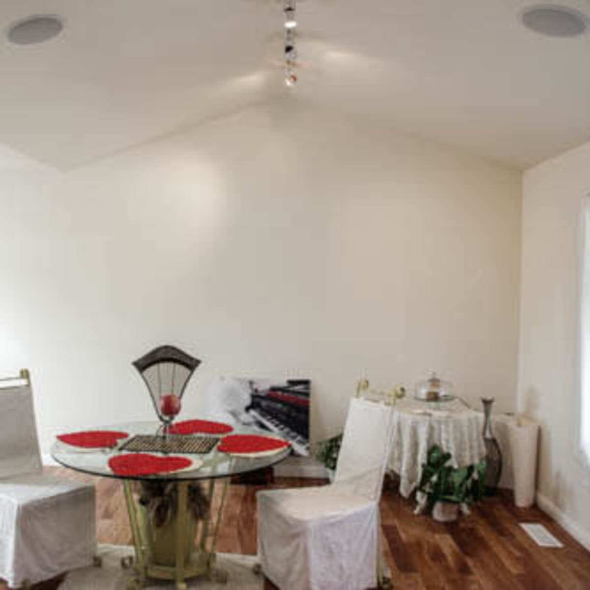 A dining area features a round table with red placemats and two chairs, surrounded by light-colored walls and hardwood flooring.