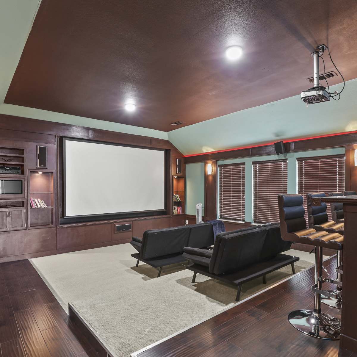 A home theater with a large screen at the front, seating arranged in two black chairs, and a bar area on one side.