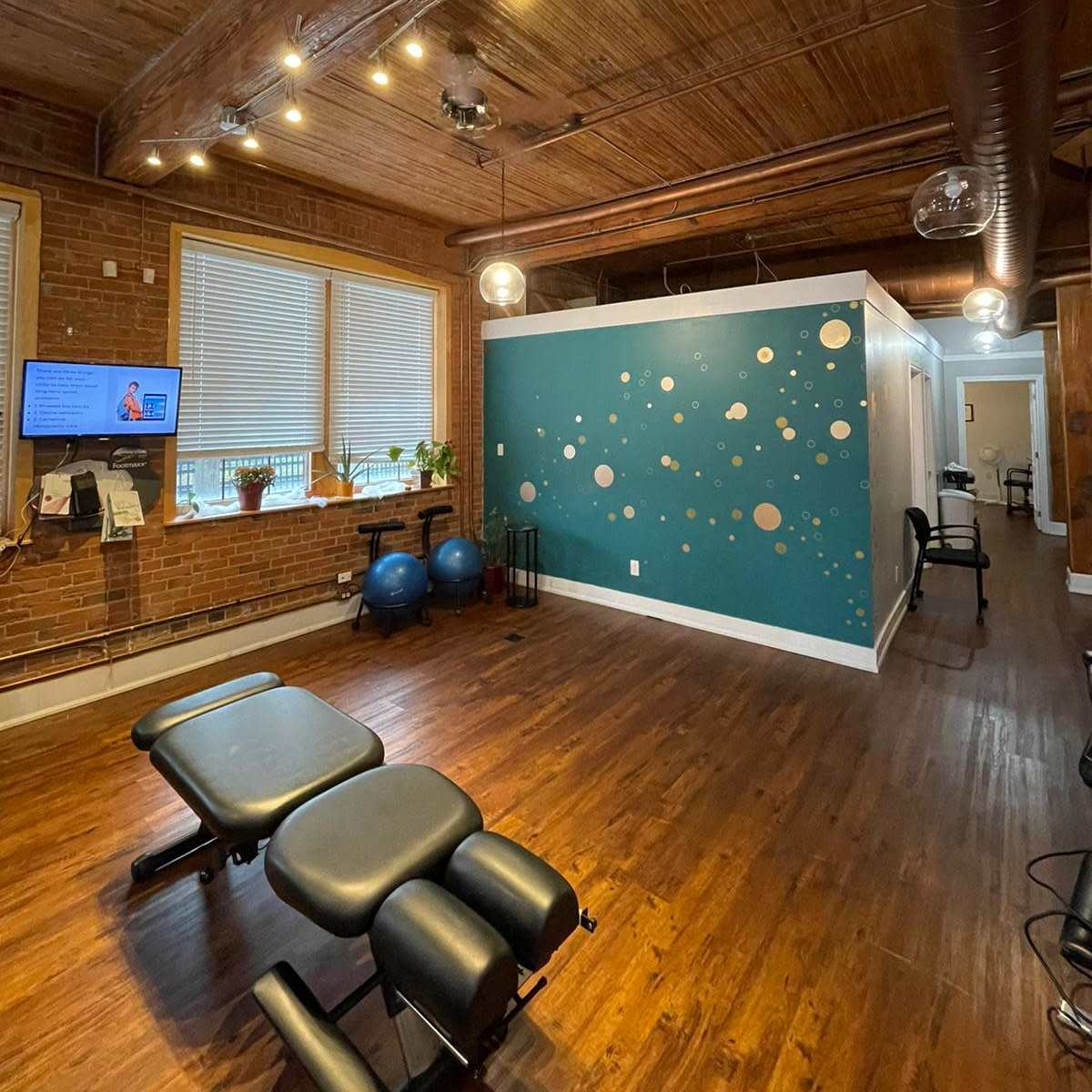 A well-lit therapy room with wooden floors, a dark green accent wall featuring circular designs, and several pieces of exercise equipment.