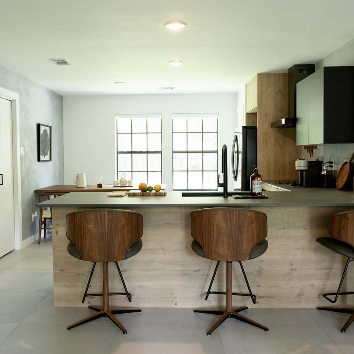 A modern kitchen features a large island with three bar stools, a dining table, and large windows that provide natural light.