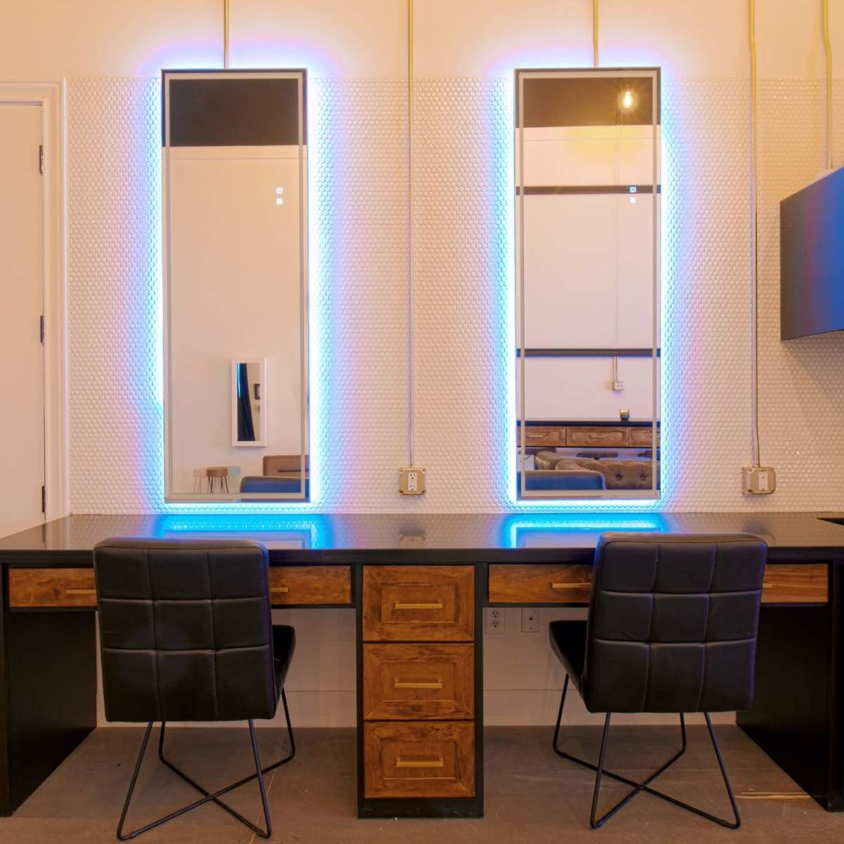 A modern vanity area with two illuminated mirrors above a sleek black desk and two black chairs, set against a textured wall.