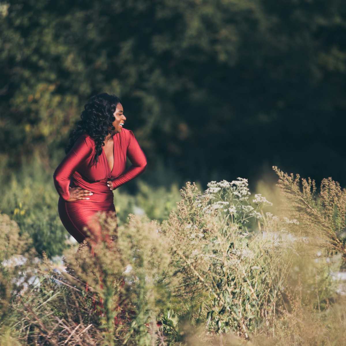 A woman in a red dress stands in a field surrounded by tall grass and wildflowers.