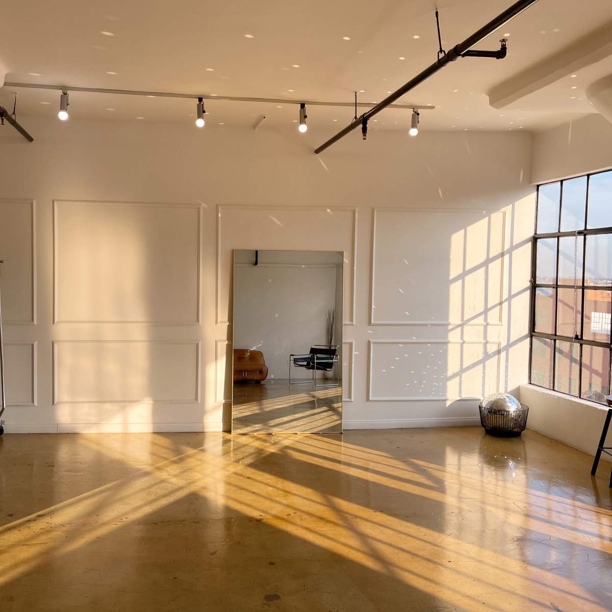 A bright room features a large window, a mirror, a stool, and a clothes rack, with sunlight casting shadows on the polished floor.