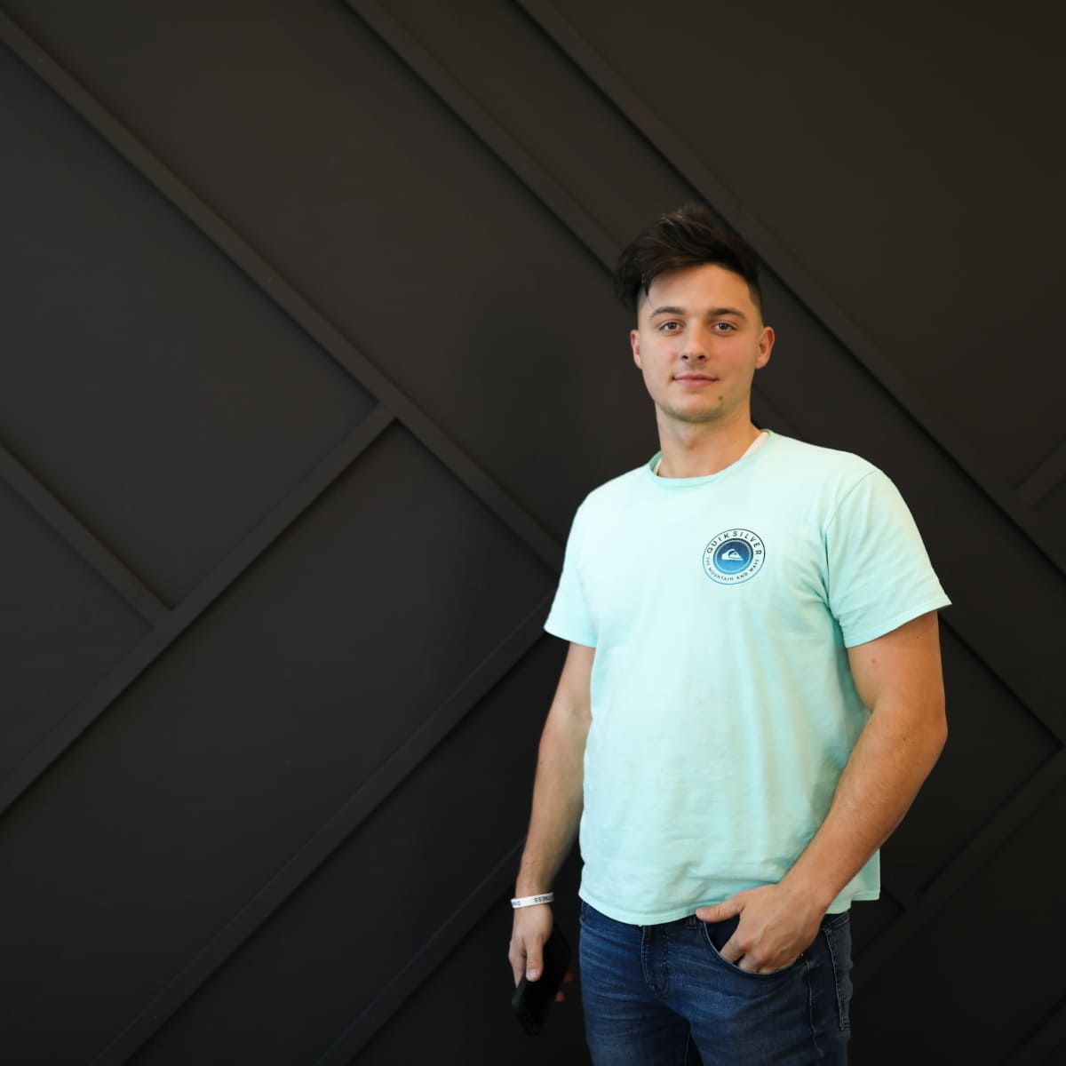 A young man stands against a dark geometric wall while wearing a light blue t-shirt and jeans.