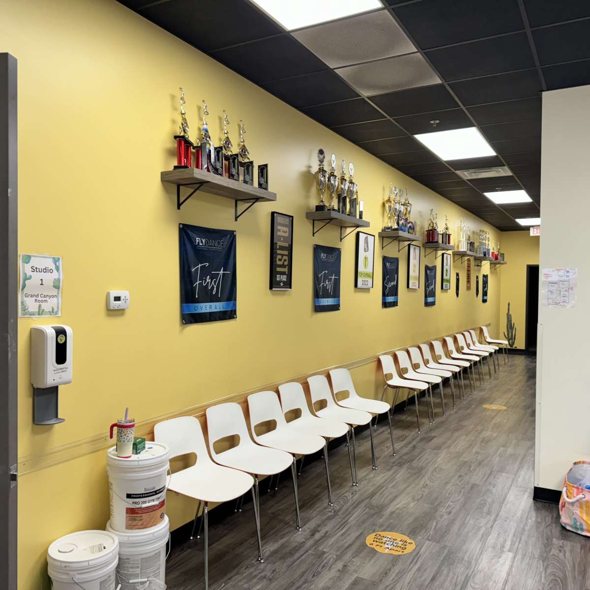 A long hallway with yellow walls, lined with chairs on one side and shelves displaying trophies and framed posters on the other.