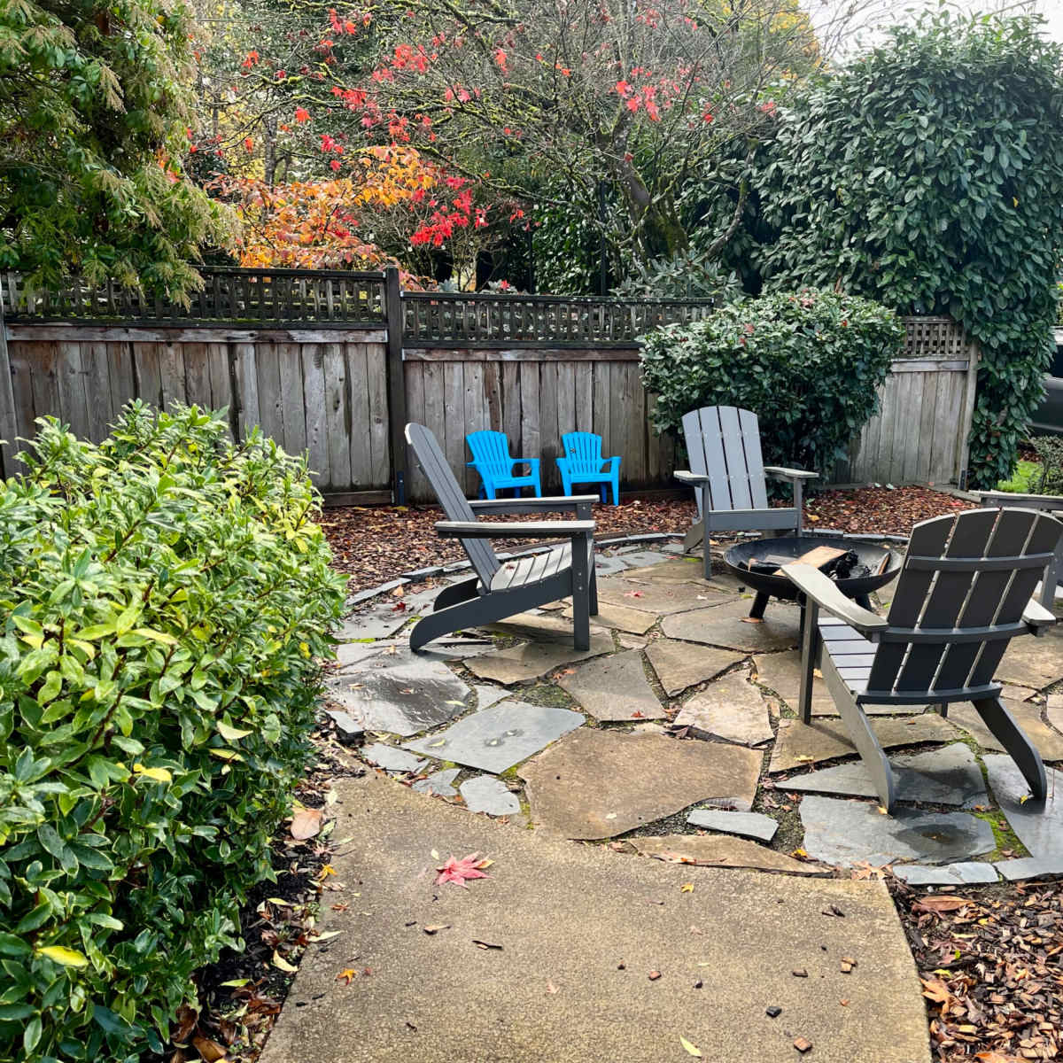 A circular stone patio surrounded by green shrubs, featuring four dark gray Adirondack chairs and two blue chairs.