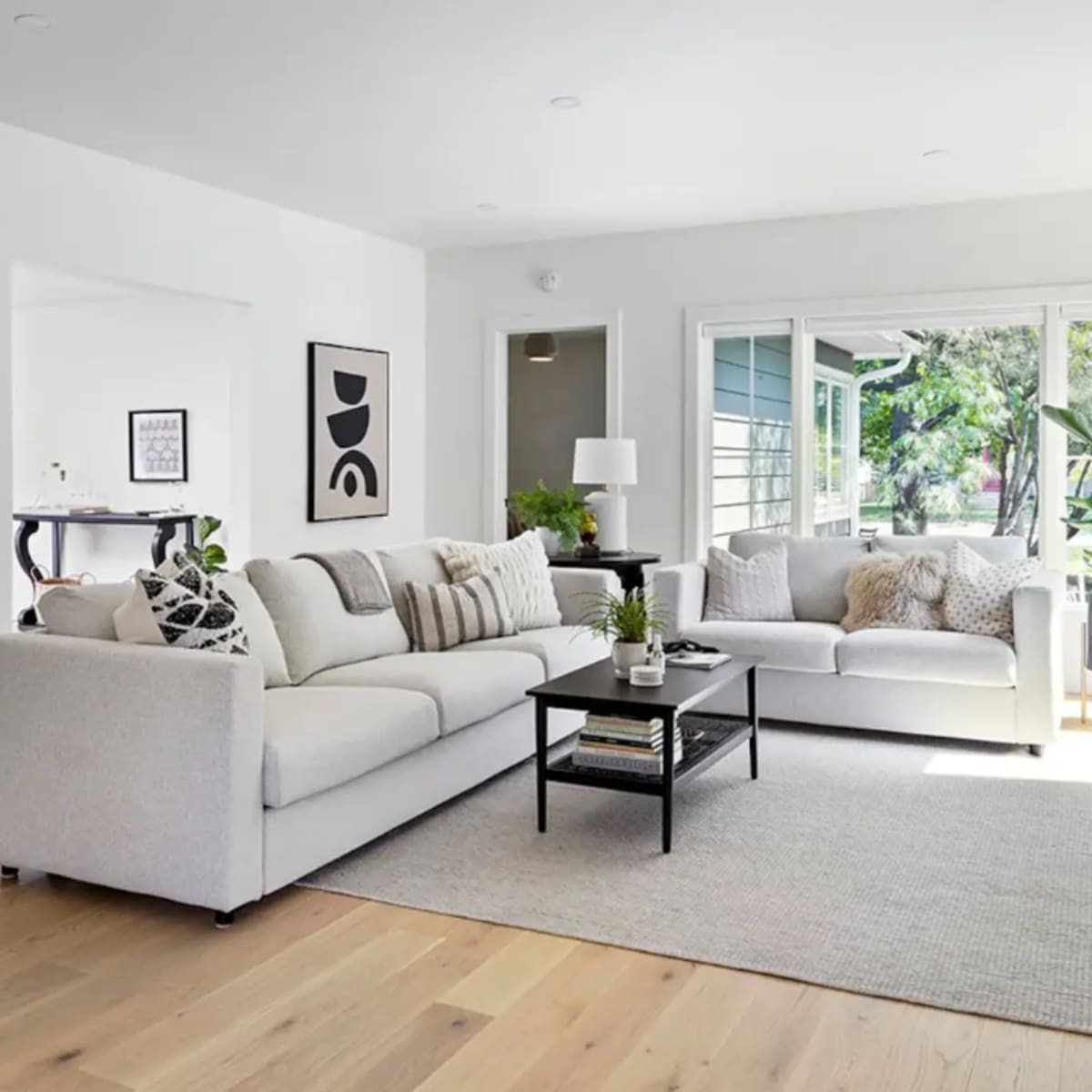 A spacious living room features a large light-colored sectional sofa, a black coffee table, and large windows allowing natural light to illuminate the space.
