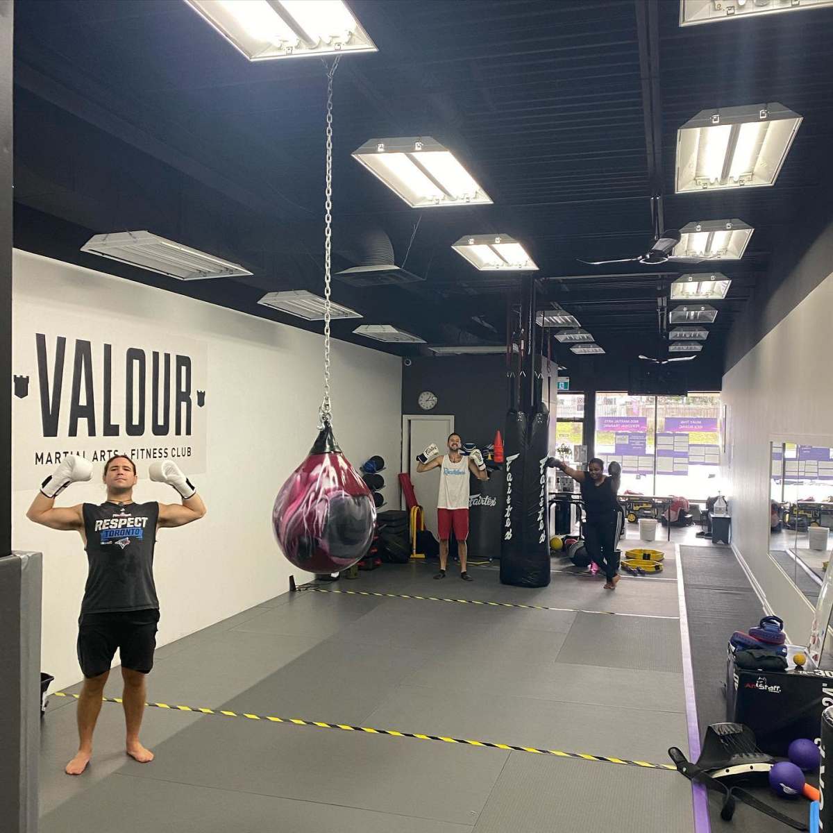 A fitness club interior features a training area with a heavy bag hanging, individuals exercising, and equipment displayed along the walls.