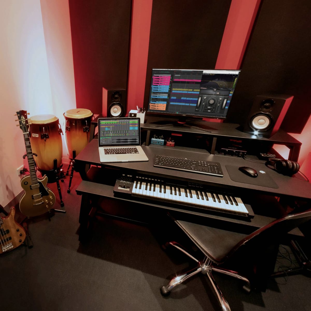 A compact music studio features a computer workstation with software displayed, musical instruments including guitars and congas, and sound equipment arranged around a black desk.