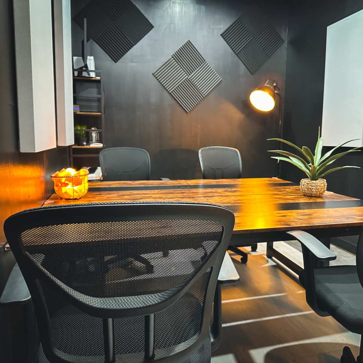 A conference room features a wooden table surrounded by ergonomic chairs, with wall-mounted acoustic panels and a lamp illuminating a small potted plant.