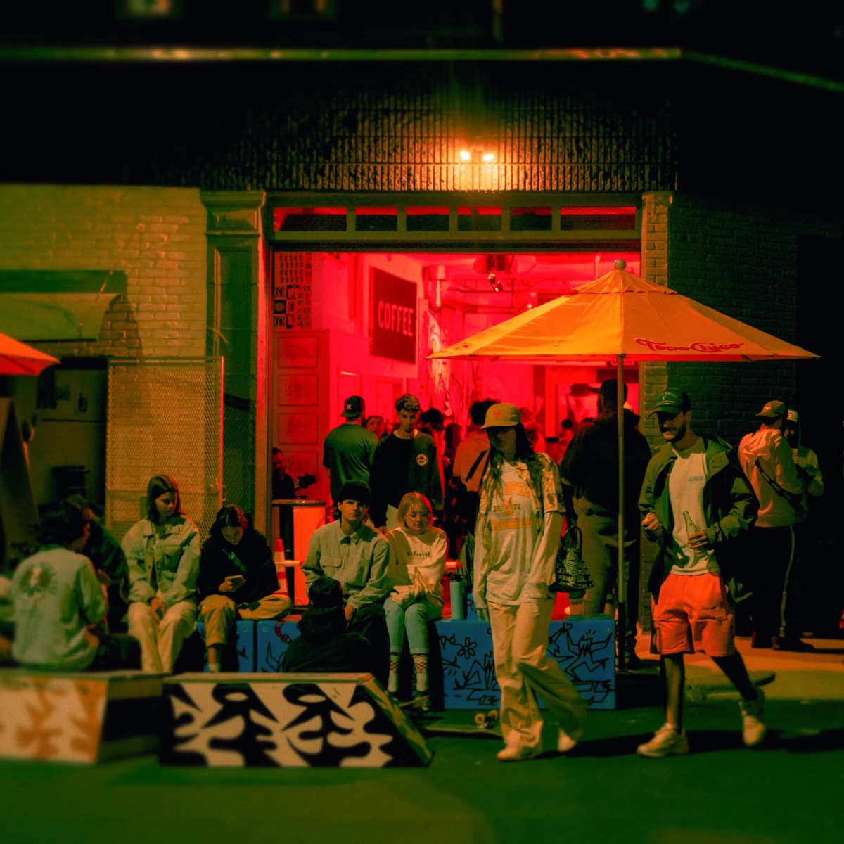 A group of people socializes outside a brightly lit coffee shop at night, with colorful umbrellas providing shade over seated patrons.