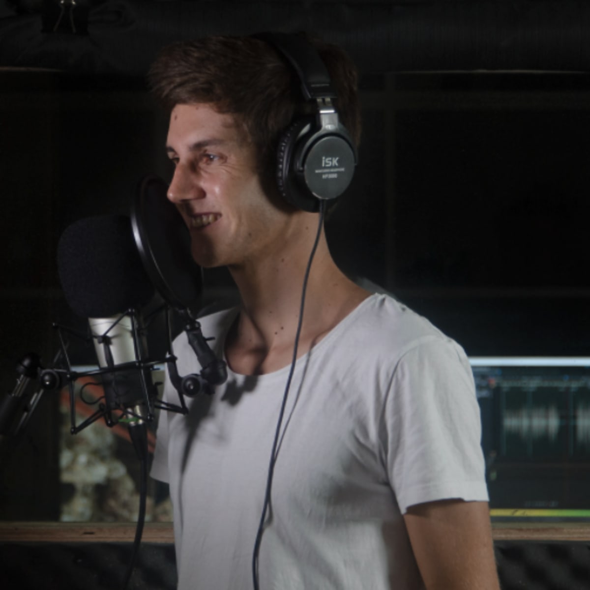 A young man wearing headphones stands in a recording studio, speaking into a microphone.