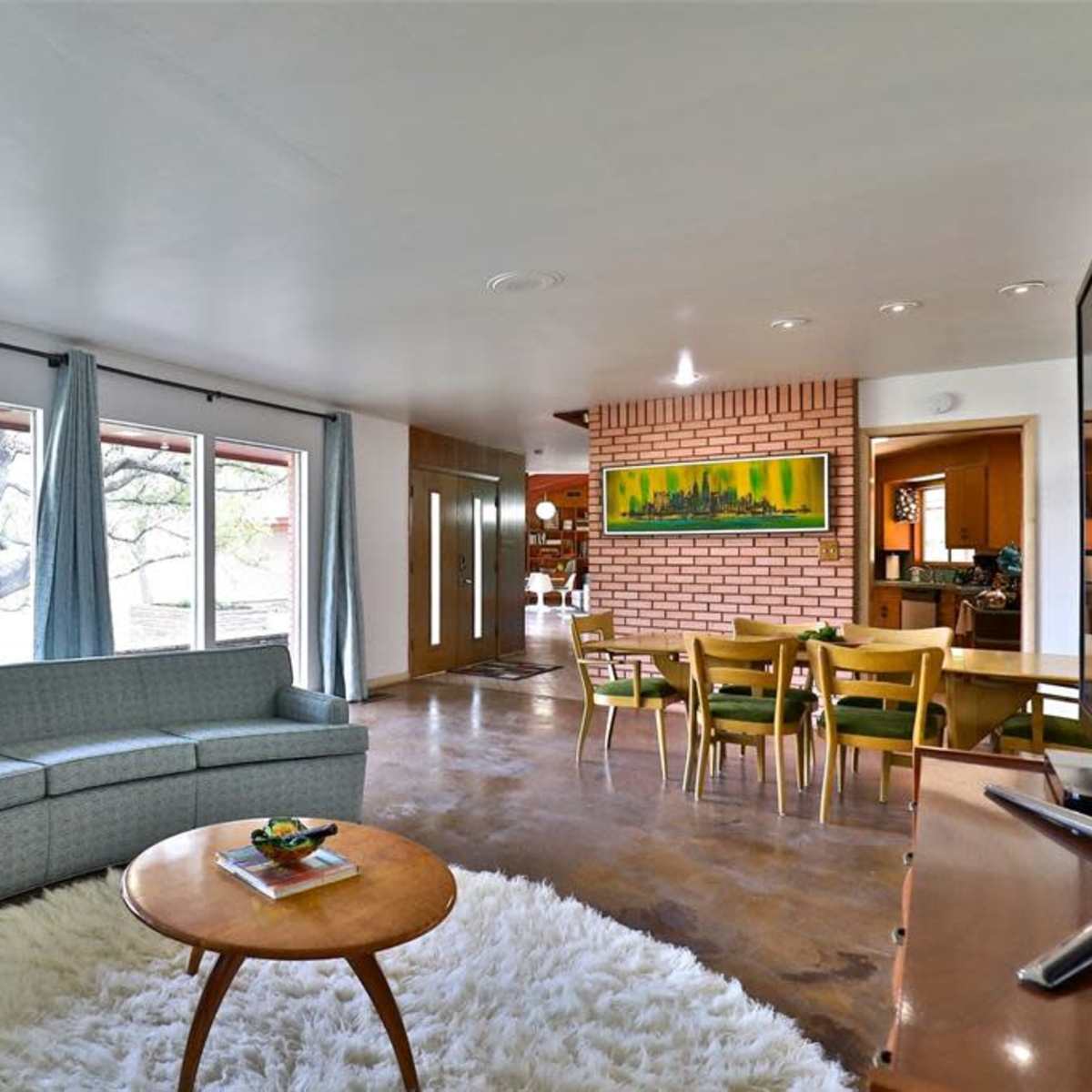 A modern living room with a curved gray sofa, a round coffee table, and large windows overlooking a tree, connected to a dining area featuring a brick wall.