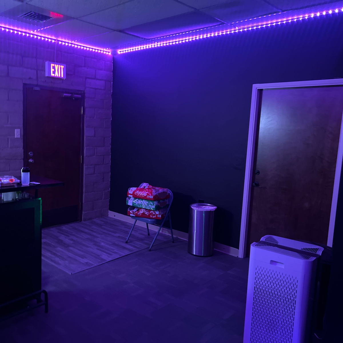 A dimly lit room with purple LED lights along the ceiling, featuring a reception desk, two doors, a stack of blankets on a chair, and a trash can.