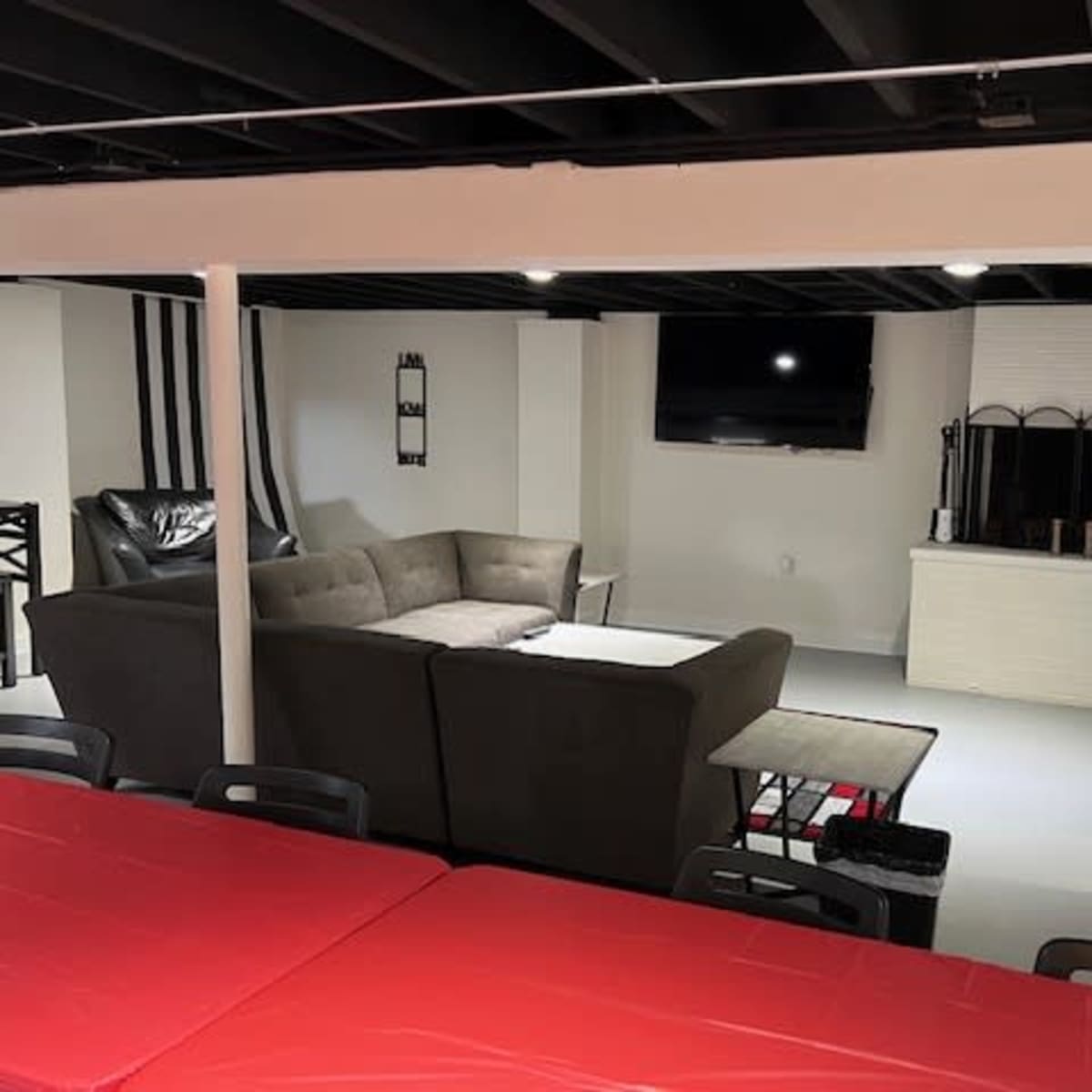 A modern basement living area with a sectional sofa, a television mounted on the wall, and a dining table with red tablecloths, along with barstools at a counter.
