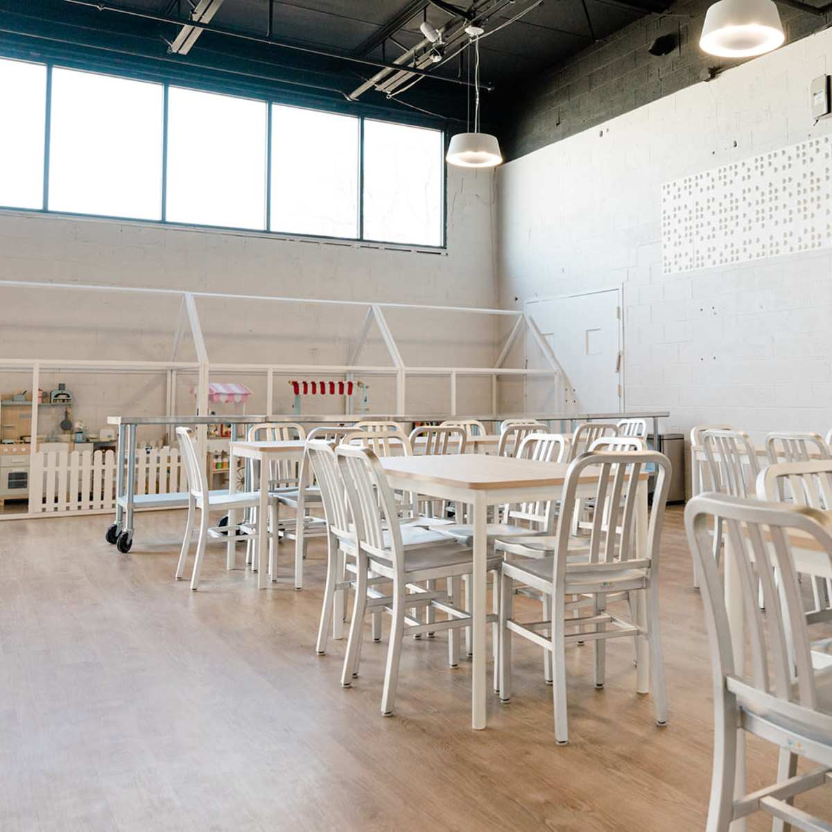A bright, spacious room with wooden tables and metal chairs arranged in rows, along with large windows letting in natural light.