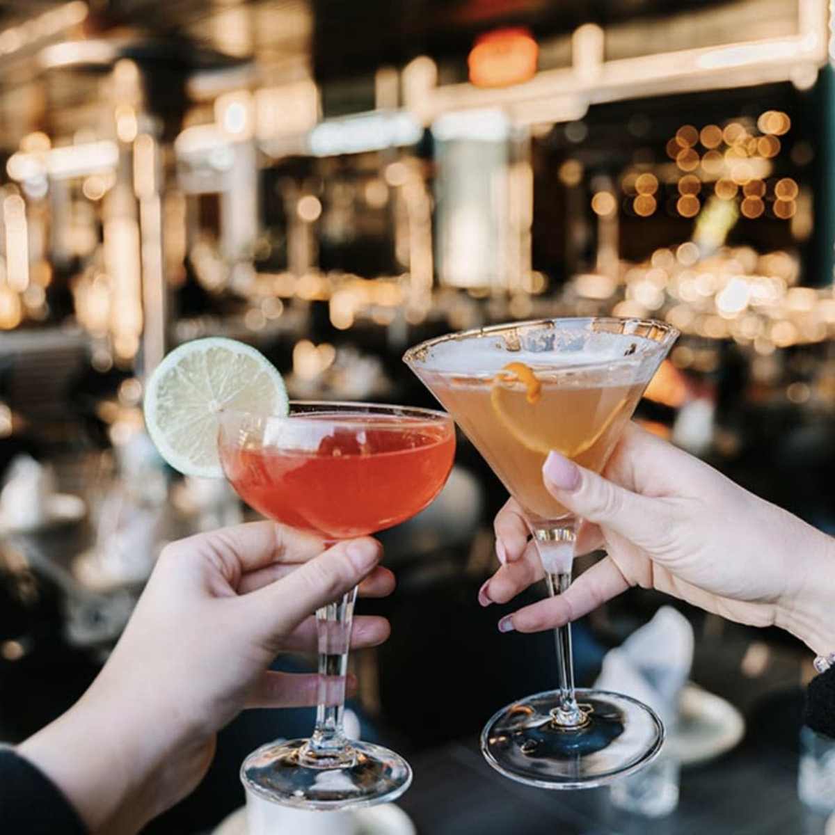 Two cocktails, one garnished with a lime and the other with an orange twist, are being clinked together in a bar setting with blurred lights in the background.