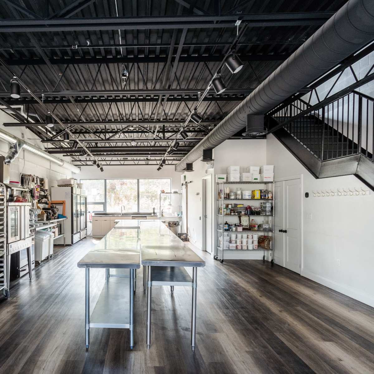 A modern commercial kitchen with stainless steel tables, various kitchen equipment, and a staircase leading to an upper level.