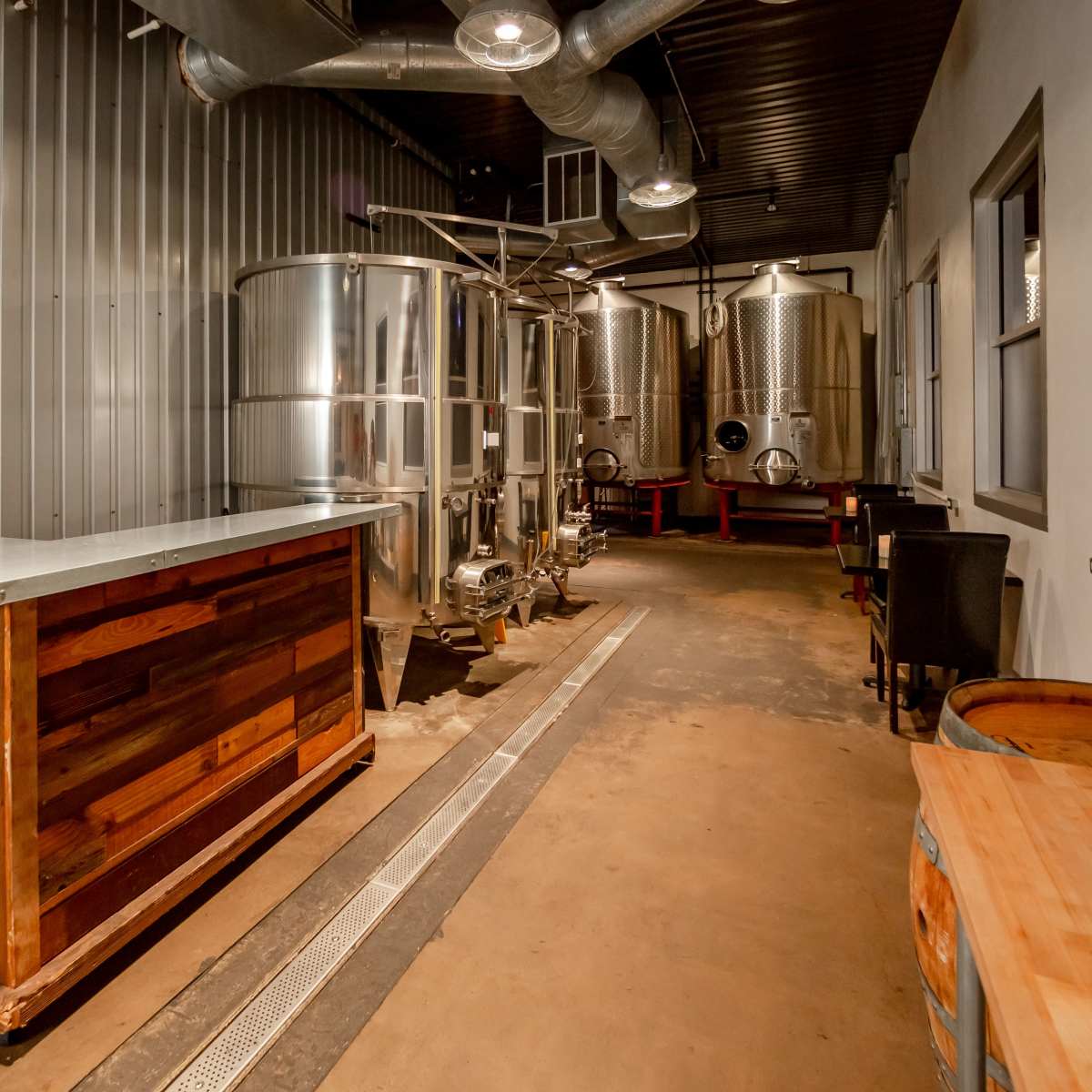 A modern brewing facility with stainless steel fermentation tanks, a wooden serving counter, and an unfinished concrete floor.