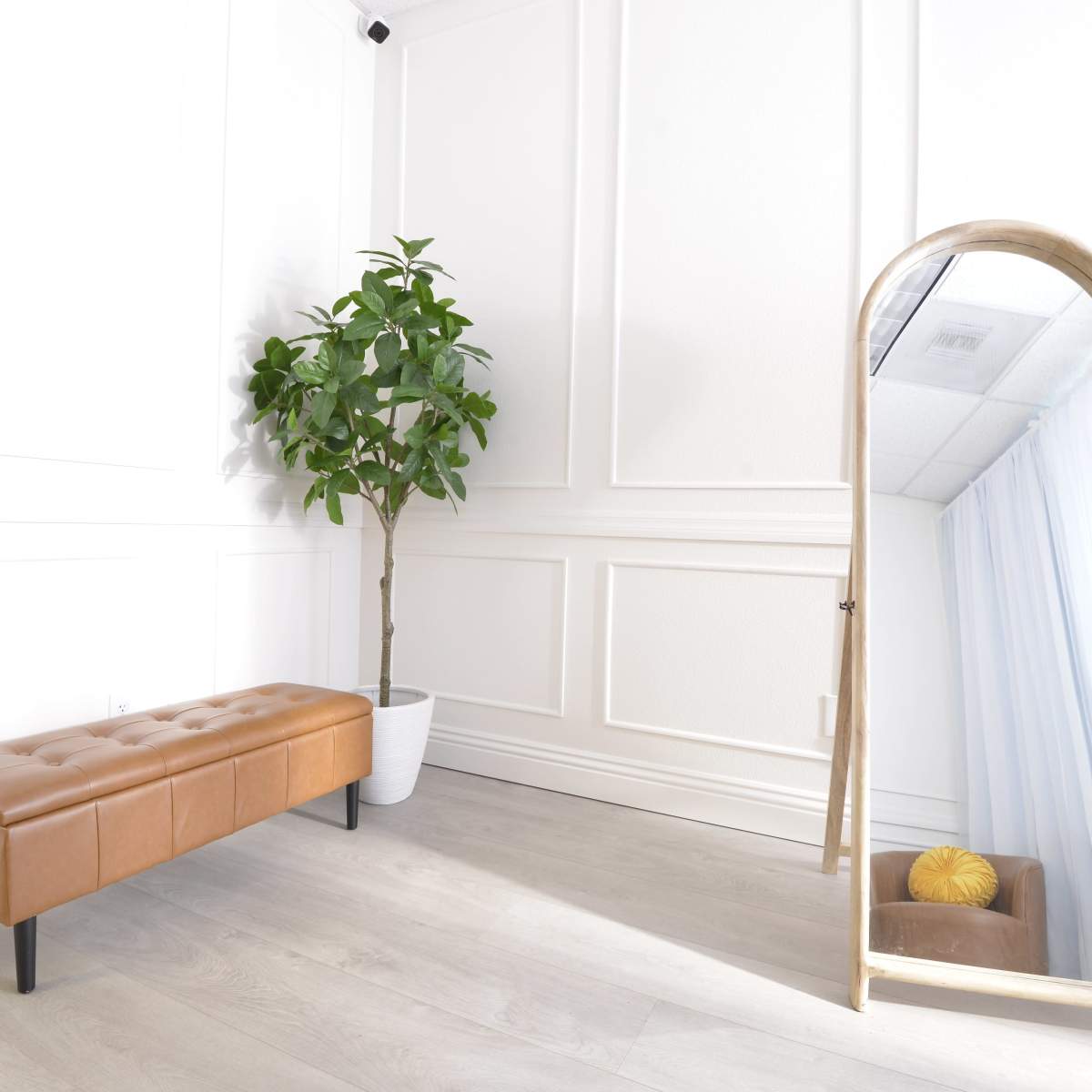 A minimalist interior with a brown upholstered bench, a potted plant in a white container, and a full-length mirror against the wall.