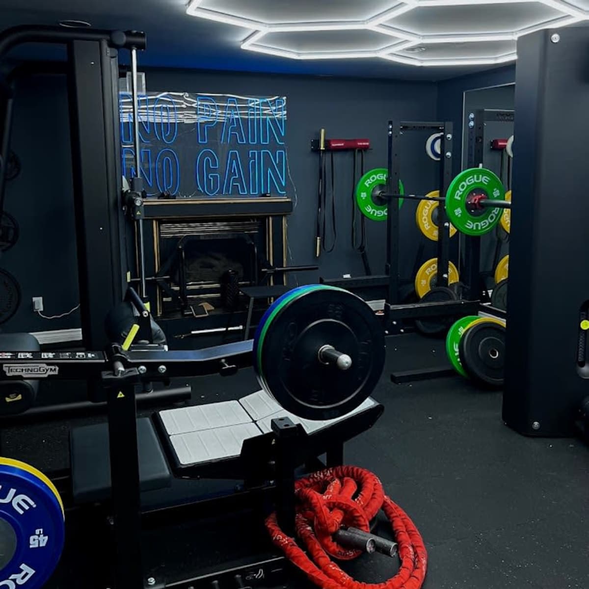 A modern gym room with various weightlifting equipment, including a bench press and colorful weight plates, along with red ropes on the floor and a motivational sign on the wall.