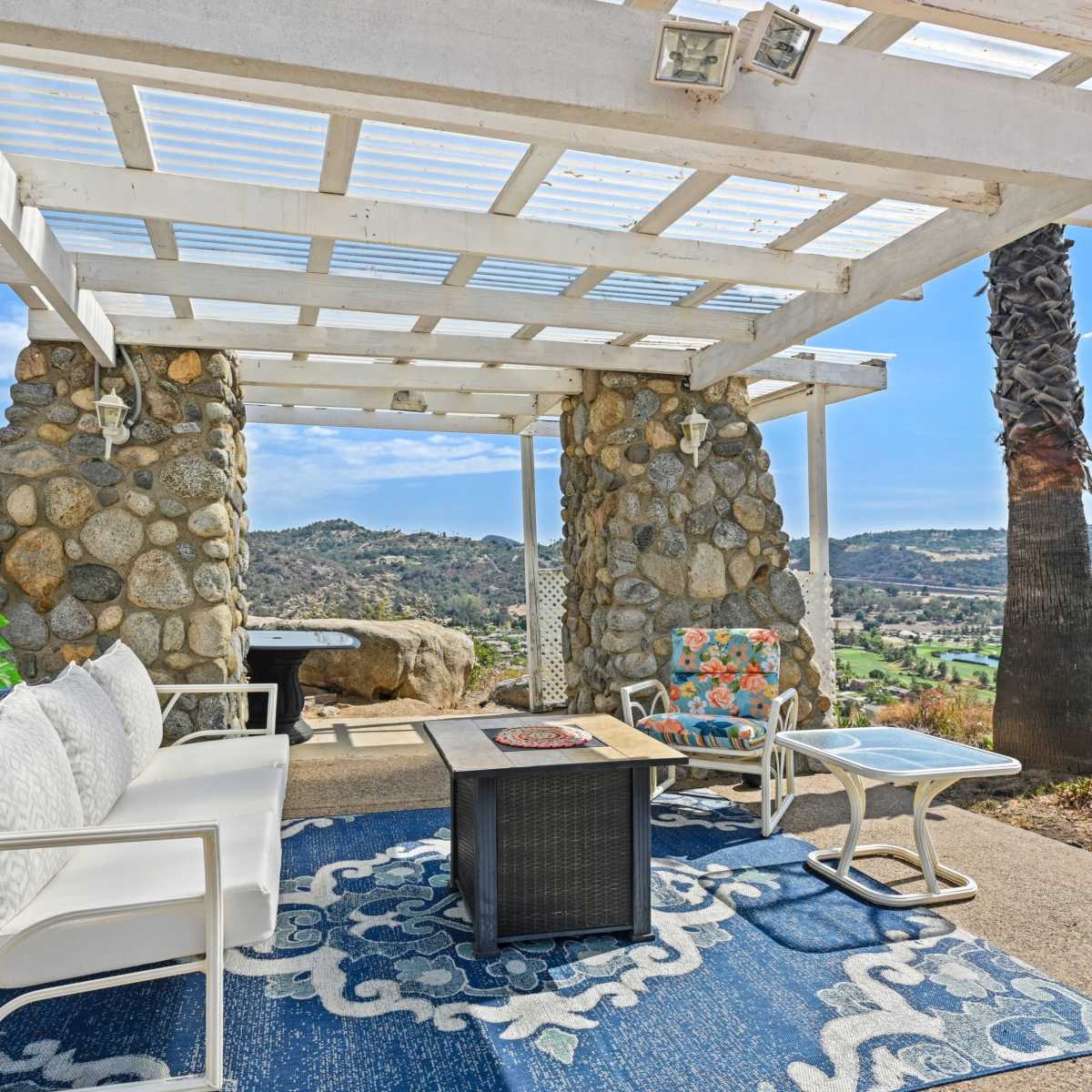 A patio area with a stone structure, white seating, and a table, set against a scenic backdrop of hills and valleys.