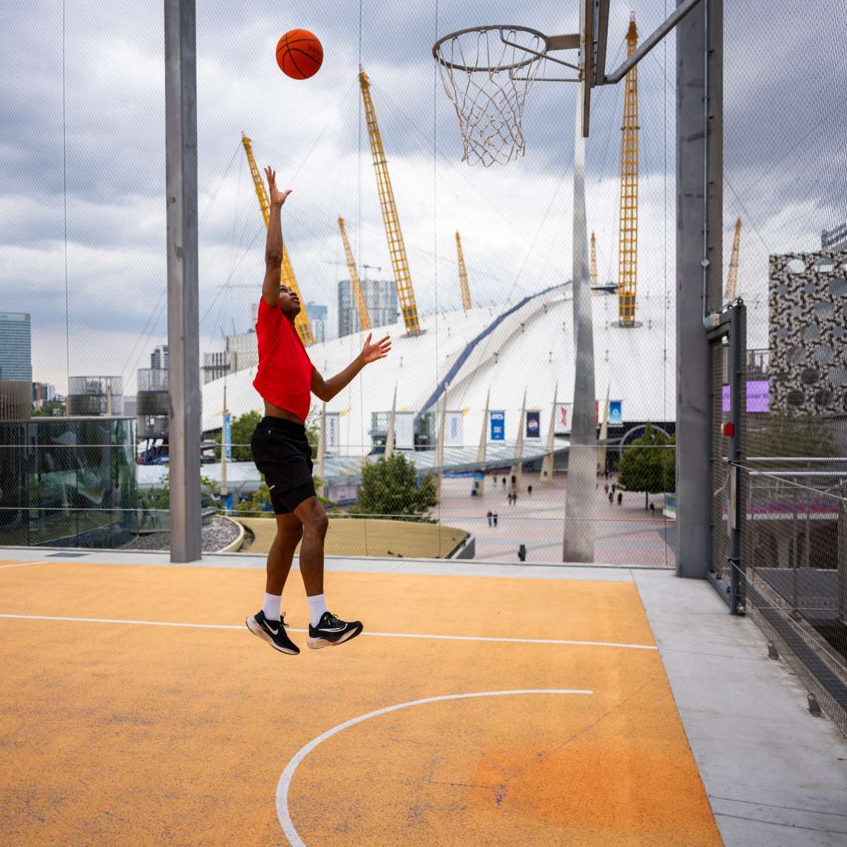 A player jumps to shoot a basketball towards a hoop on a rooftop court with a city skyline in the background.