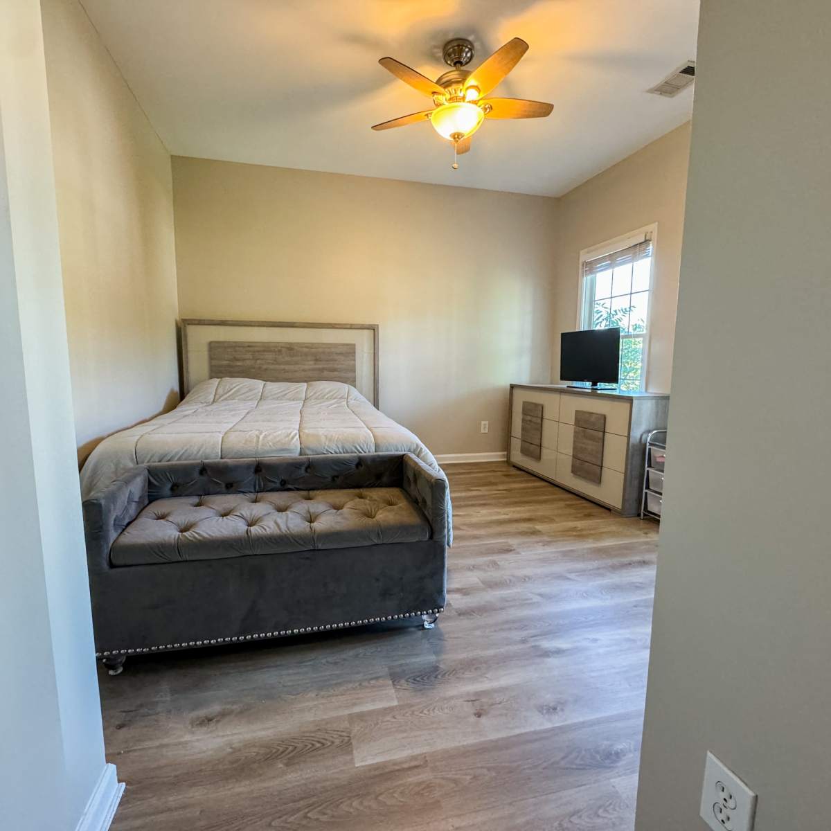 A bedroom features a gray bed frame with a tufted base, a wooden dresser, and a small television against a light-colored wall.