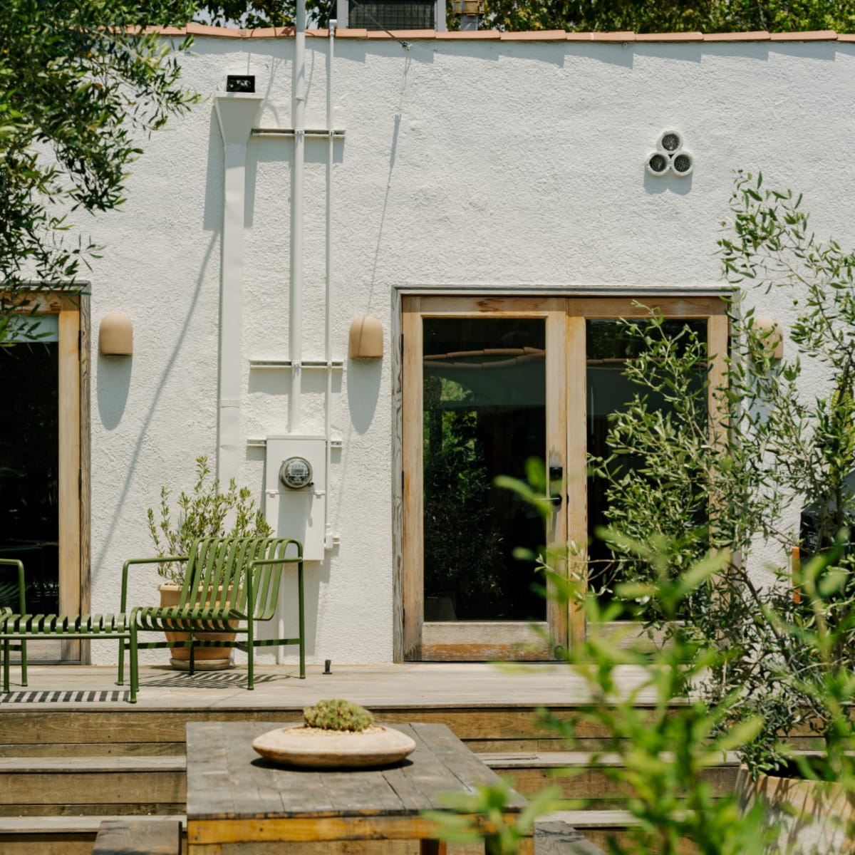 A green metal seating set is placed on a wooden deck in front of two large windows of a white stucco building.