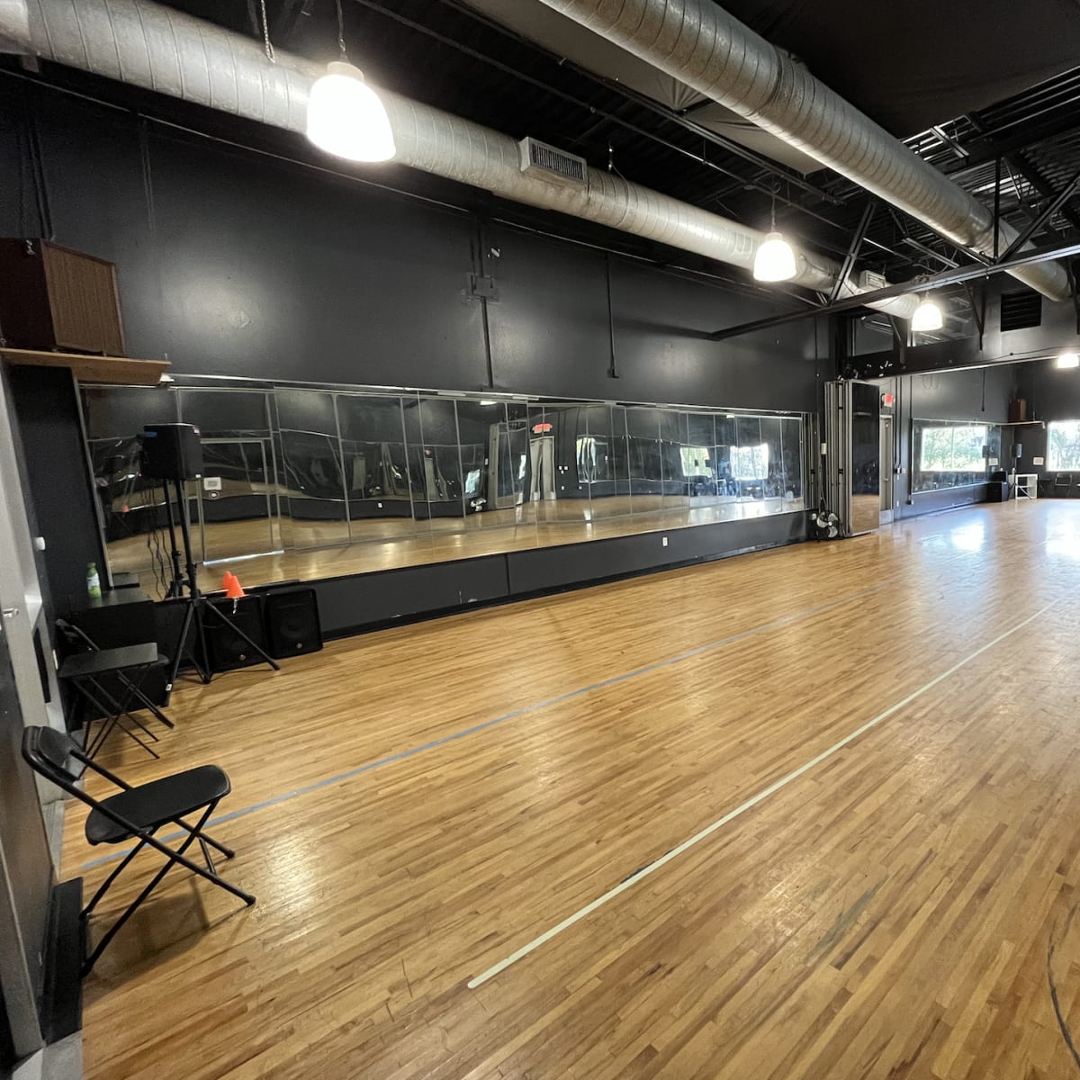 An empty dance studio with wooden floors, large mirrors on one wall, and fluorescent lights in a black-walled space.