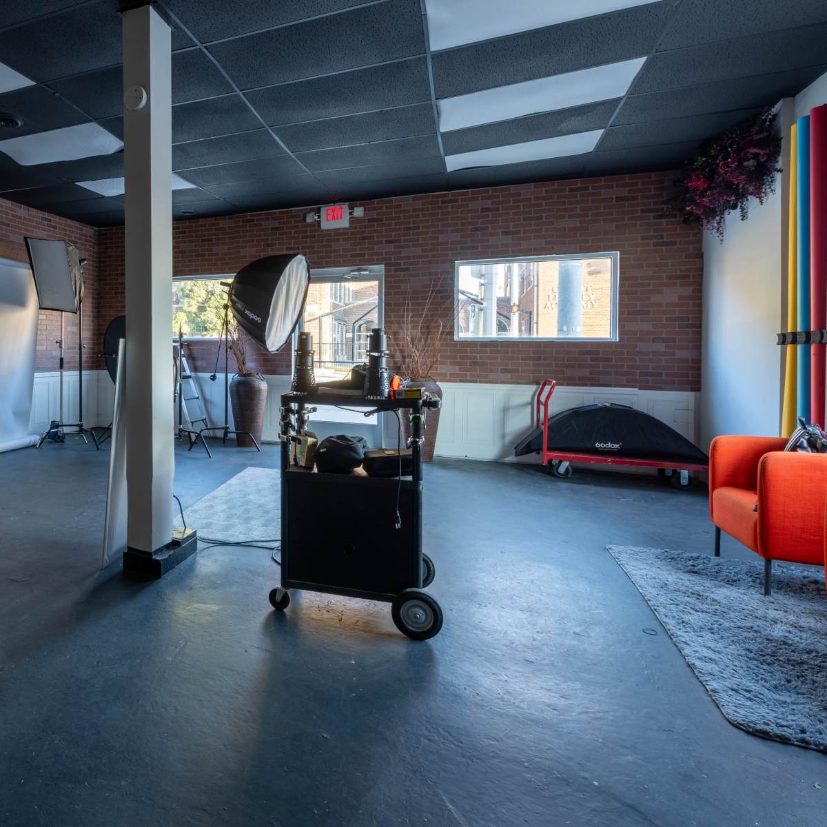 A spacious photography studio with soft lighting equipment, a backdrop stand, and colorful decorations against brick walls.