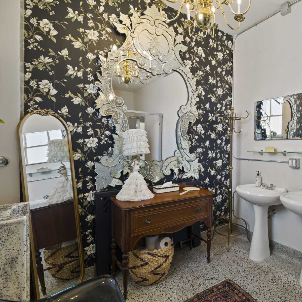 The bathroom features a floral wallpaper, a large ornate mirror, dual sinks, and a vintage-style chair next to a decorative wooden dresser.