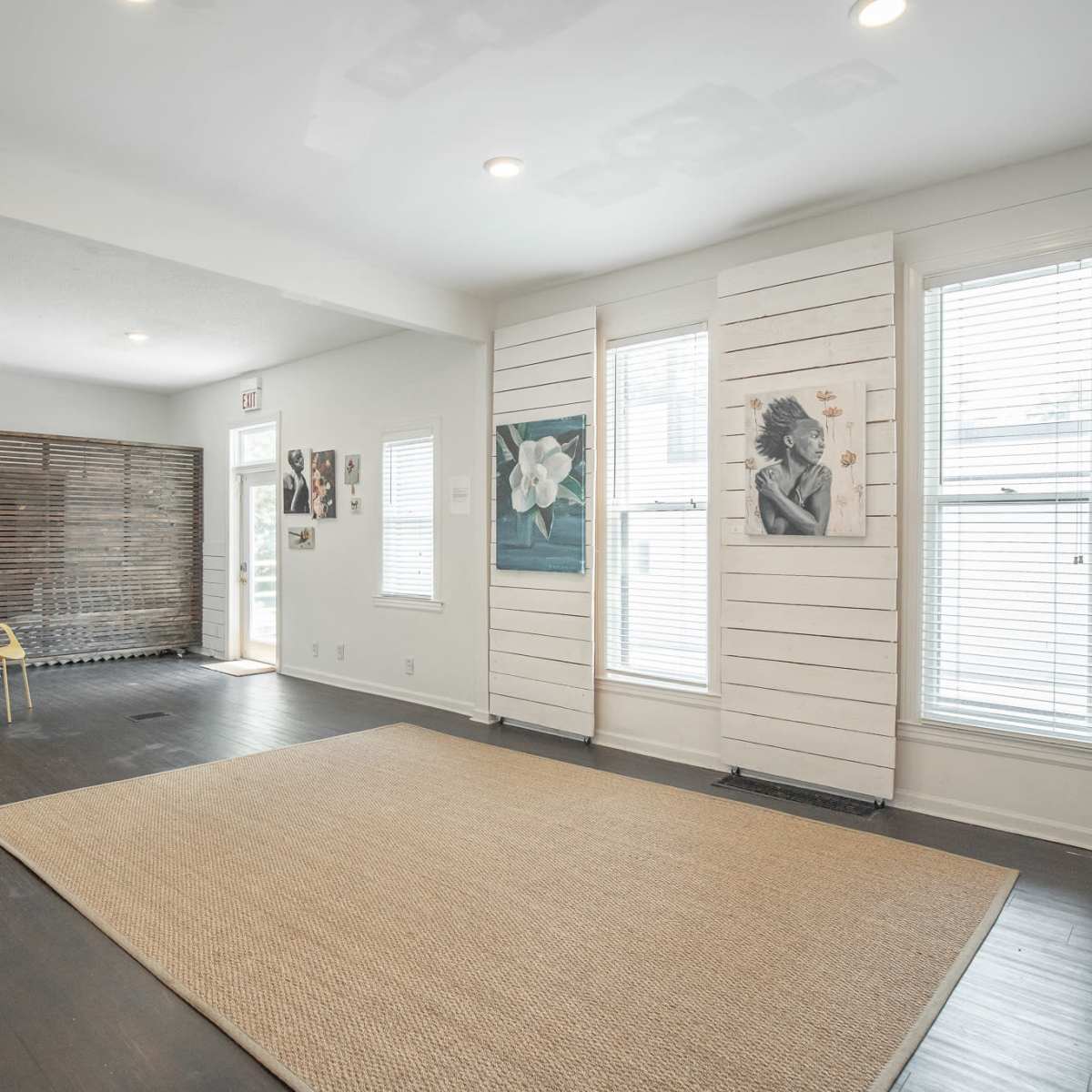 A spacious room with a large area rug, wooden floors, and natural light streaming through windows, featuring artwork on the walls and a chair in the corner.