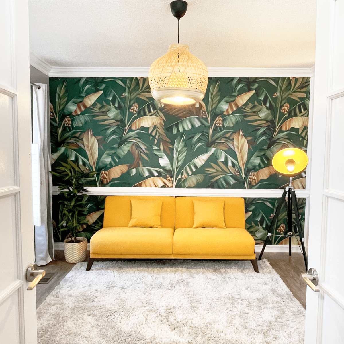 A room with a yellow sofa, a patterned green wallpaper featuring tropical leaves, and a modern lamp, viewed through glass double doors.