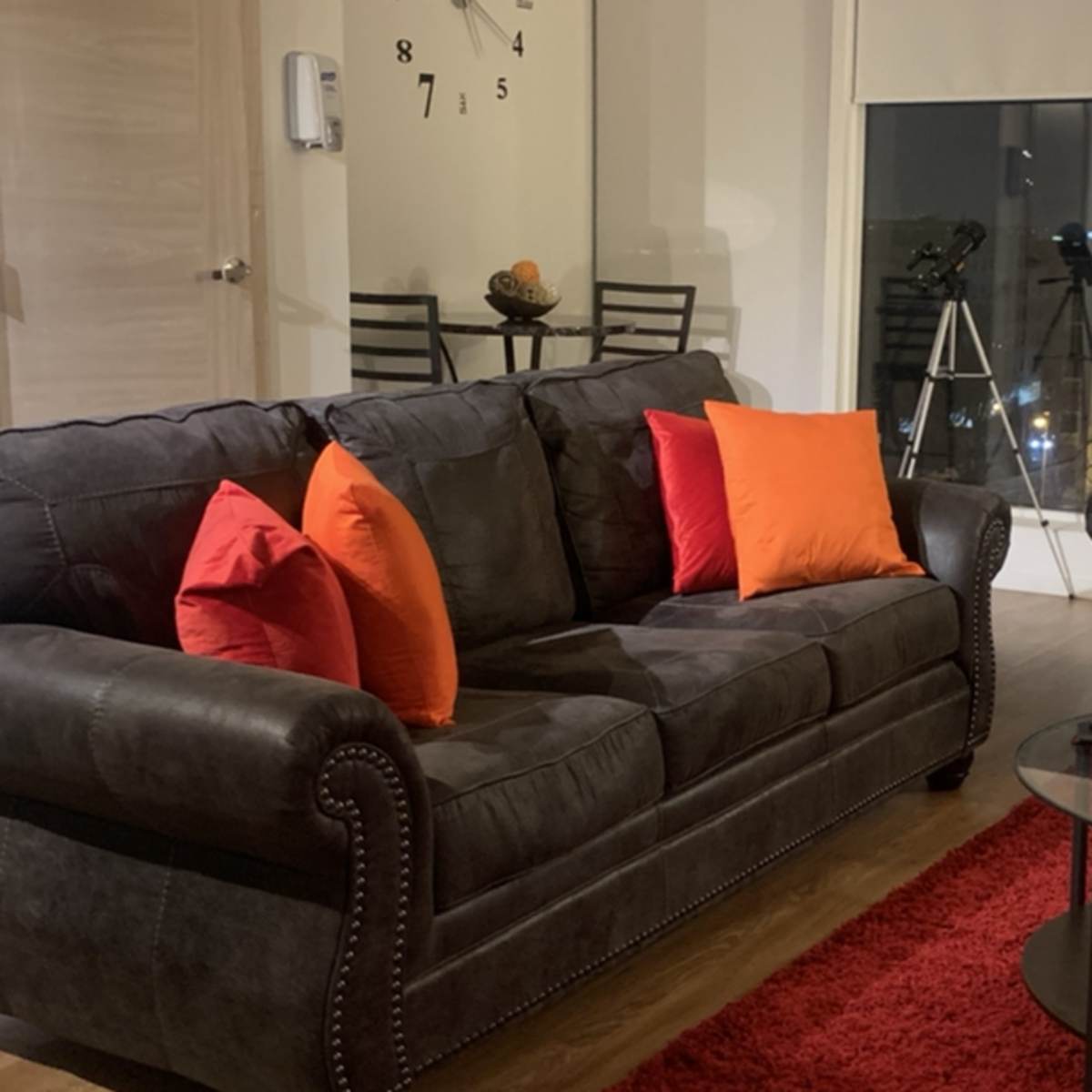 A black sofa adorned with orange pillows sits in a modern living room, complemented by a round glass coffee table and a tripod telescope in the background.