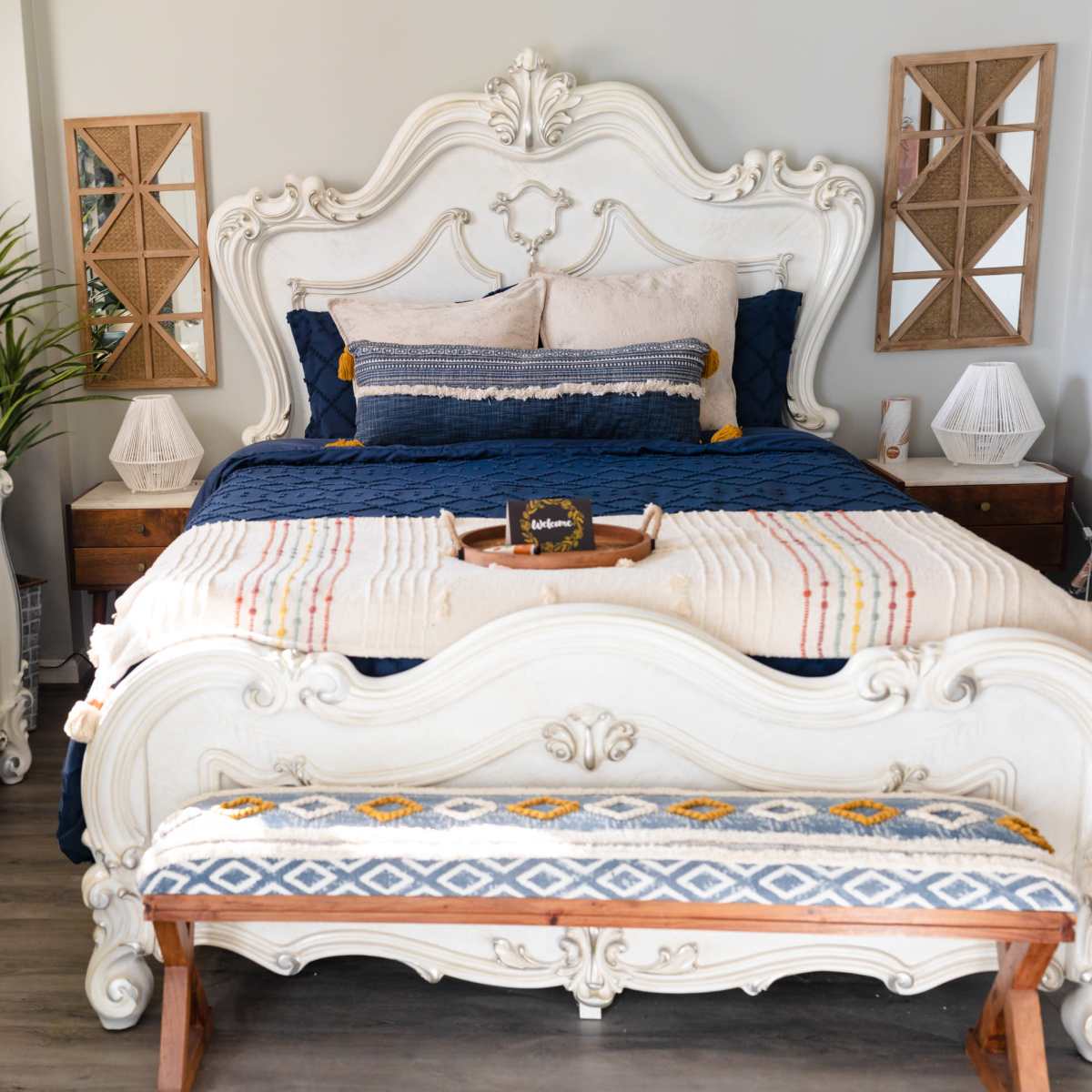 A decorated bedroom featuring a large ornate bed with blue bedding, a wooden bench at the foot, and two decorative mirrors on the walls.