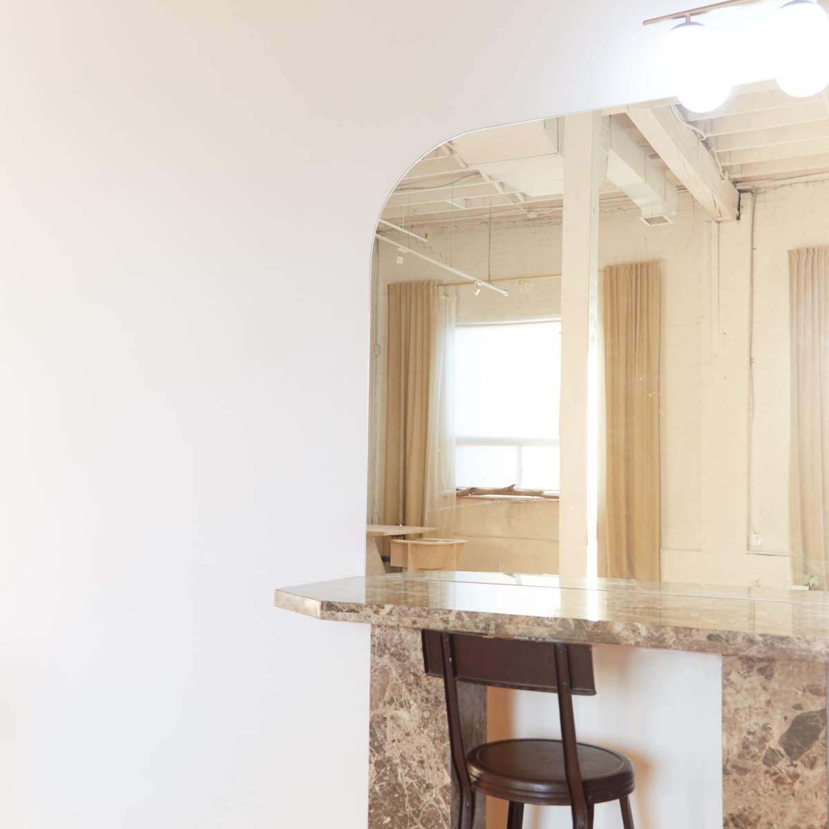 A minimalist interior features a marble countertop with a single wooden chair and a large arched mirror above it.