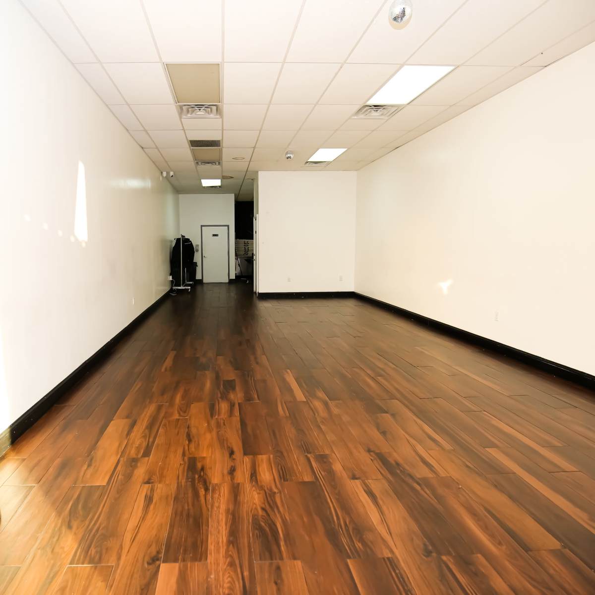 A spacious, empty room with wooden flooring and white walls, featuring a small doorway at the far end.