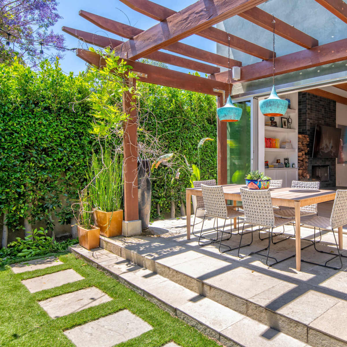 A patio area with a dining table and chairs, surrounded by greenery and sunlight.