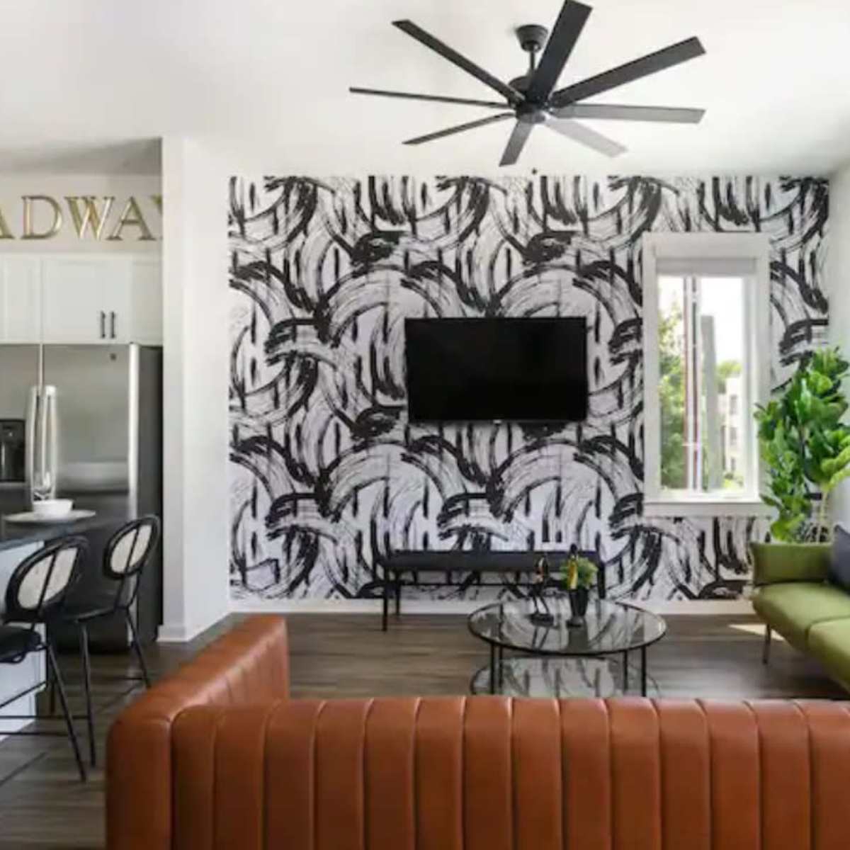 A modern living room featuring a brown leather sofa, a green couch, a black and white patterned accent wall, and a television mounted on the wall, with large windows letting in natural light.