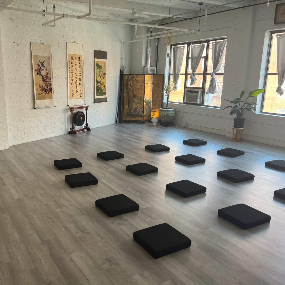 A spacious meditation room features several black cushions arranged on a light wood floor, surrounded by traditional Asian artwork and a plant.