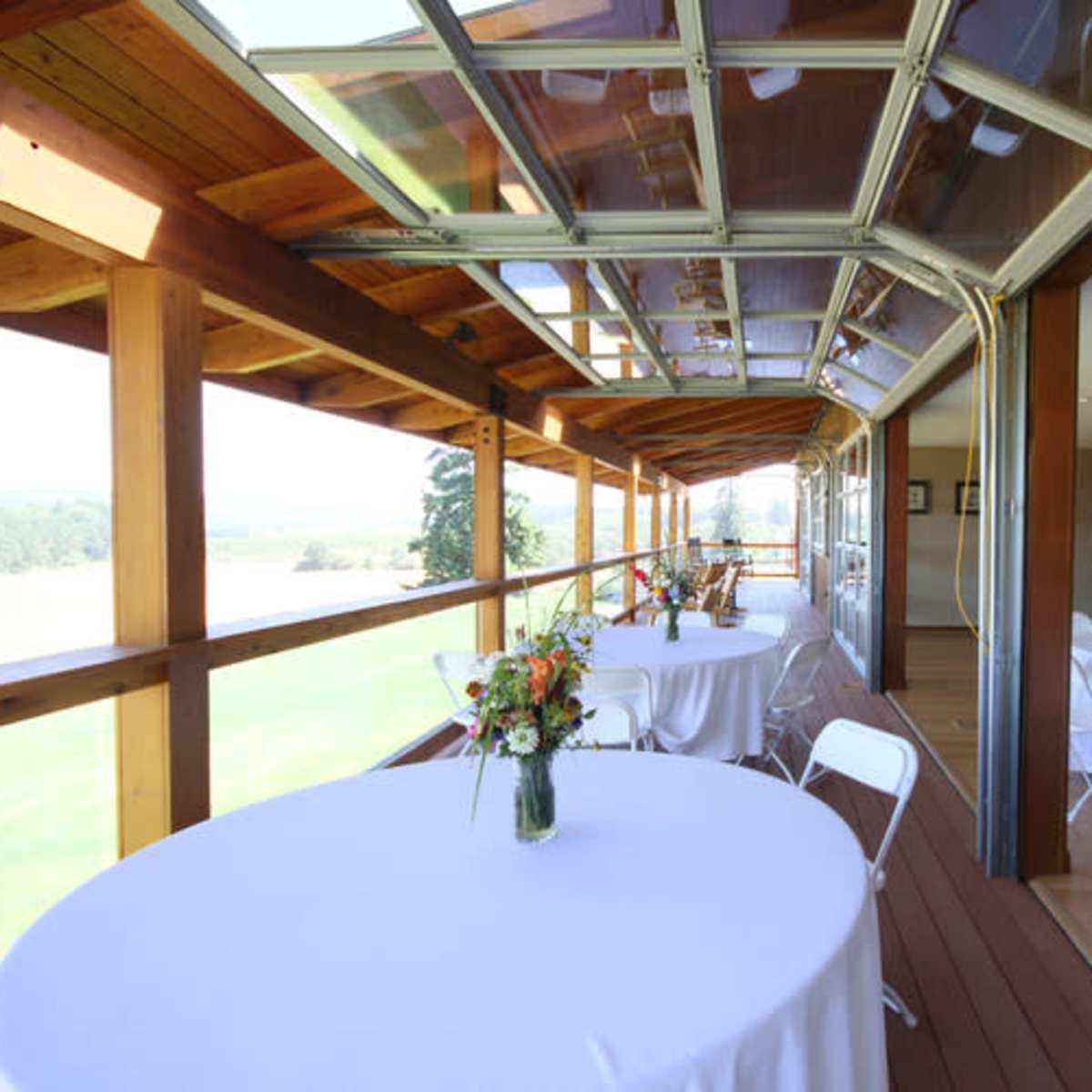 An indoor space with large glass windows, featuring white tables with floral centerpieces arranged along a wooden deck overlooking a scenic view.