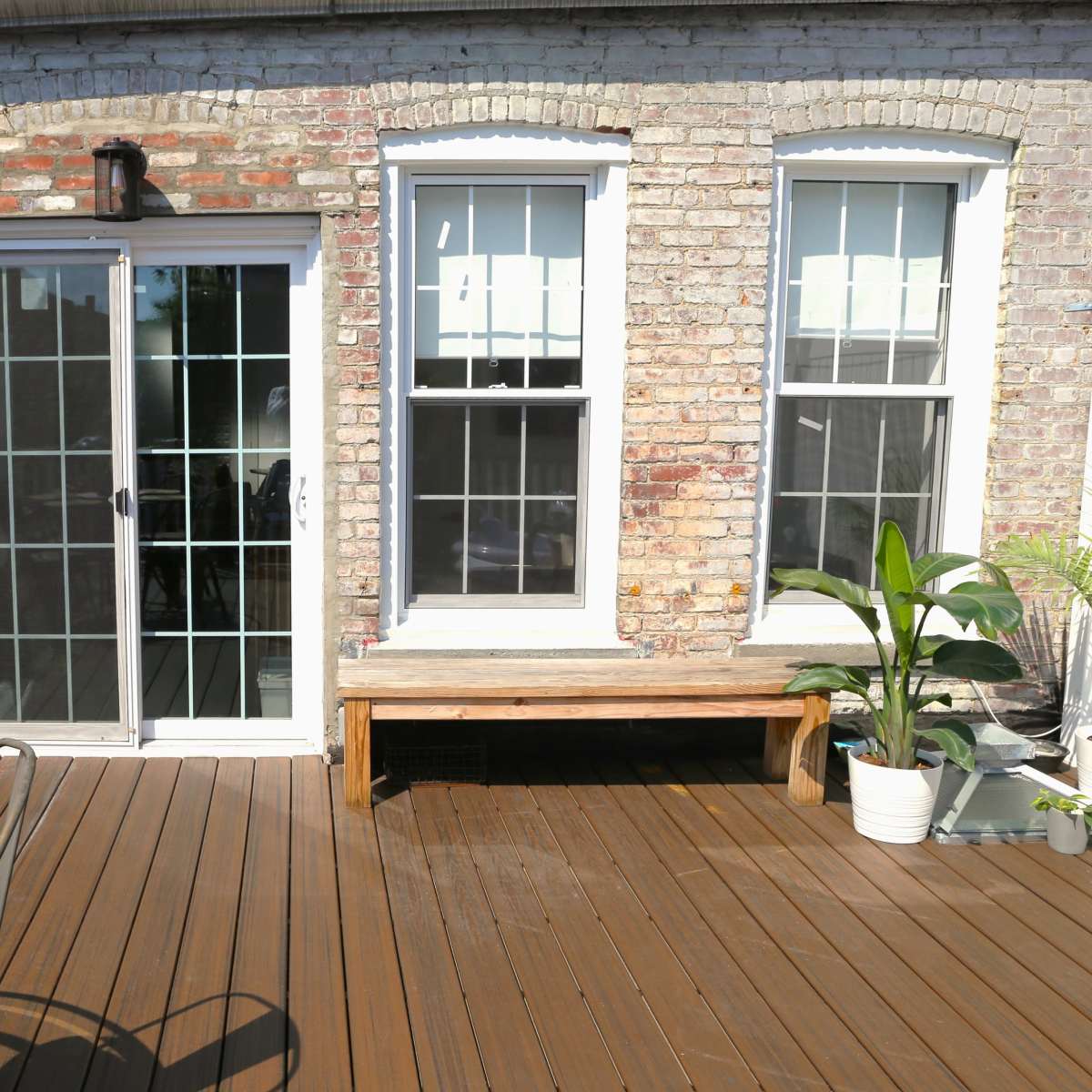 A wooden deck with a glass door leading to a brick wall house and two windows, alongside a table and potted plants.