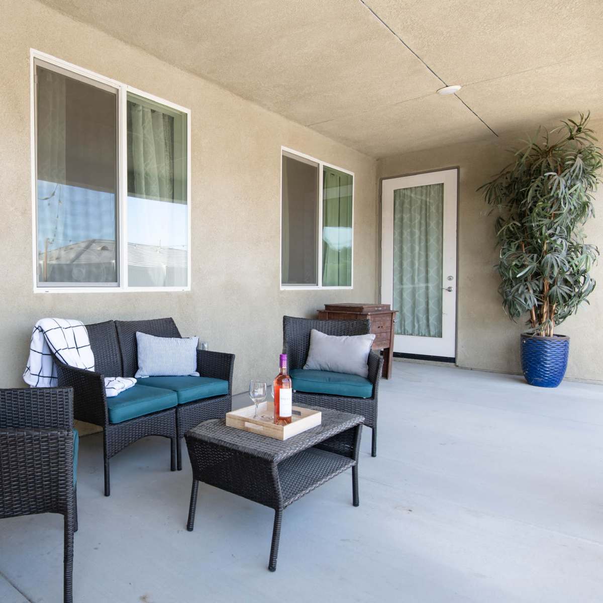 A patio area with four wicker chairs, a small table, and a large potted plant, all situated against a beige wall and large windows.