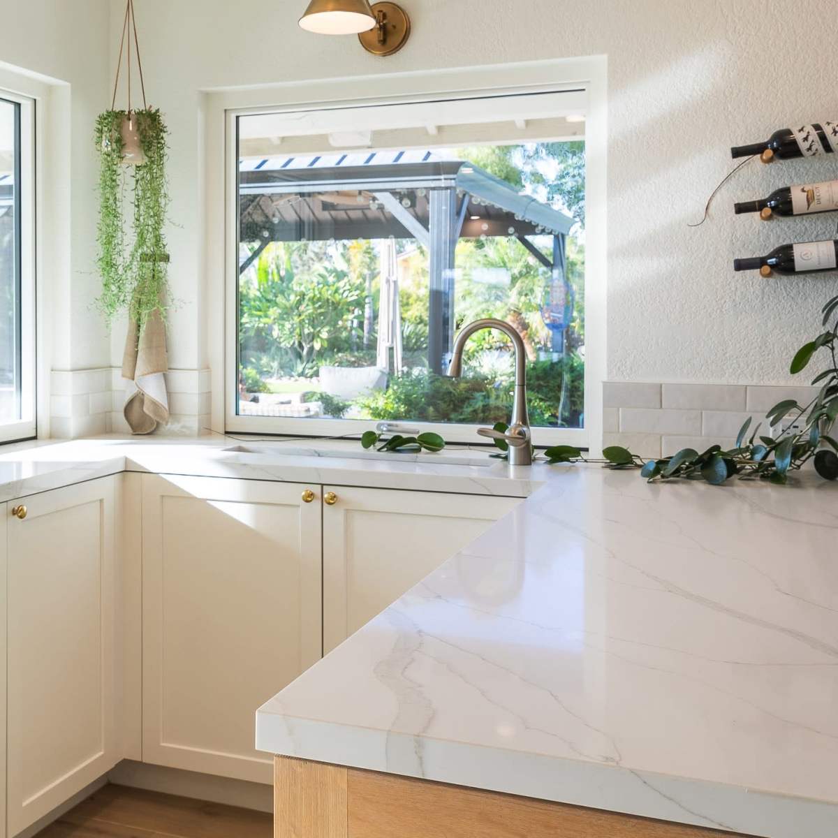 A bright kitchen features white cabinets, a marble countertop, and large windows overlooking a patio with greenery.